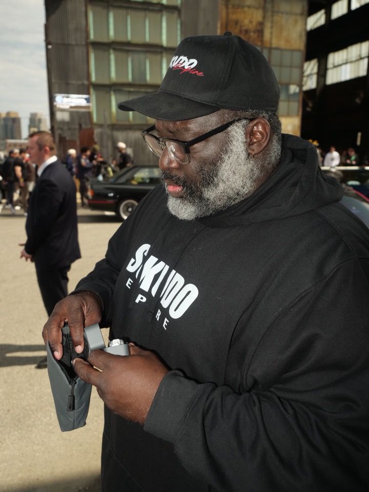 Man with gray beard and glasses wearing a black cap and hoodie, holding a smartphone outdoors near a body of water.