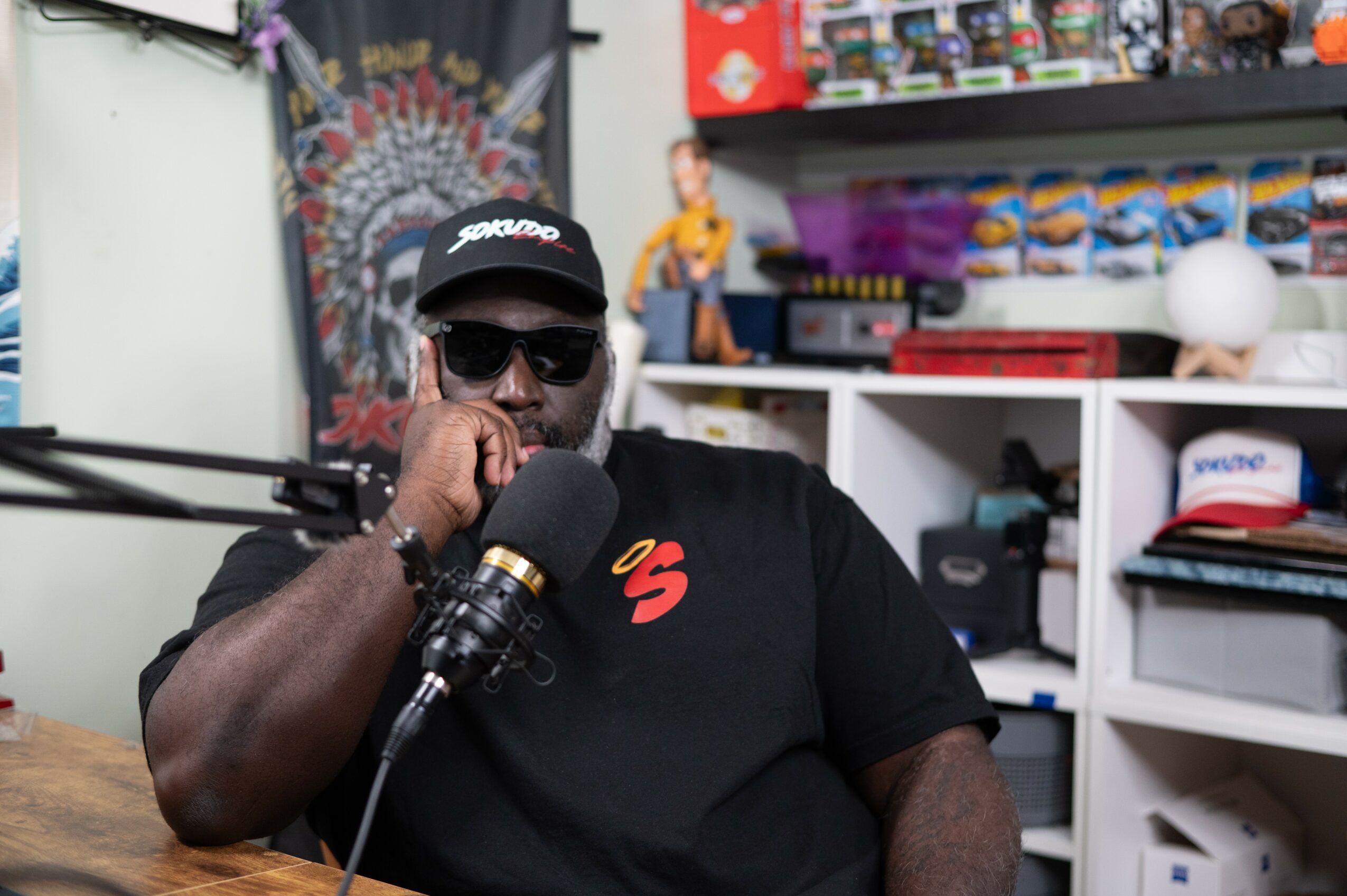Man wearing sunglasses and a cap, speaking into a microphone in a room with shelves and toys.