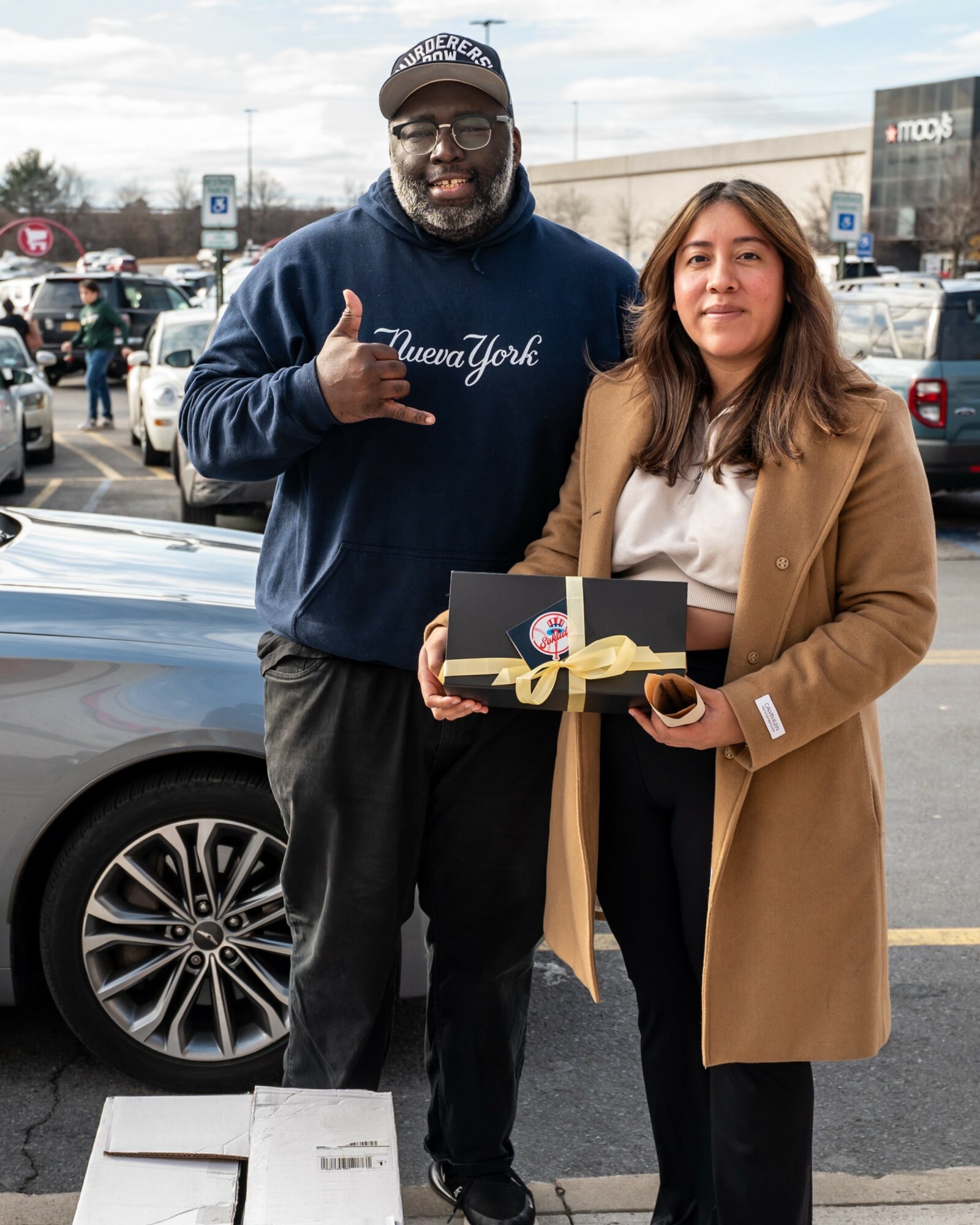 A man and woman stand outdoors in a parking lot, holding a gift box with a gold ribbon, smiling at the camera.