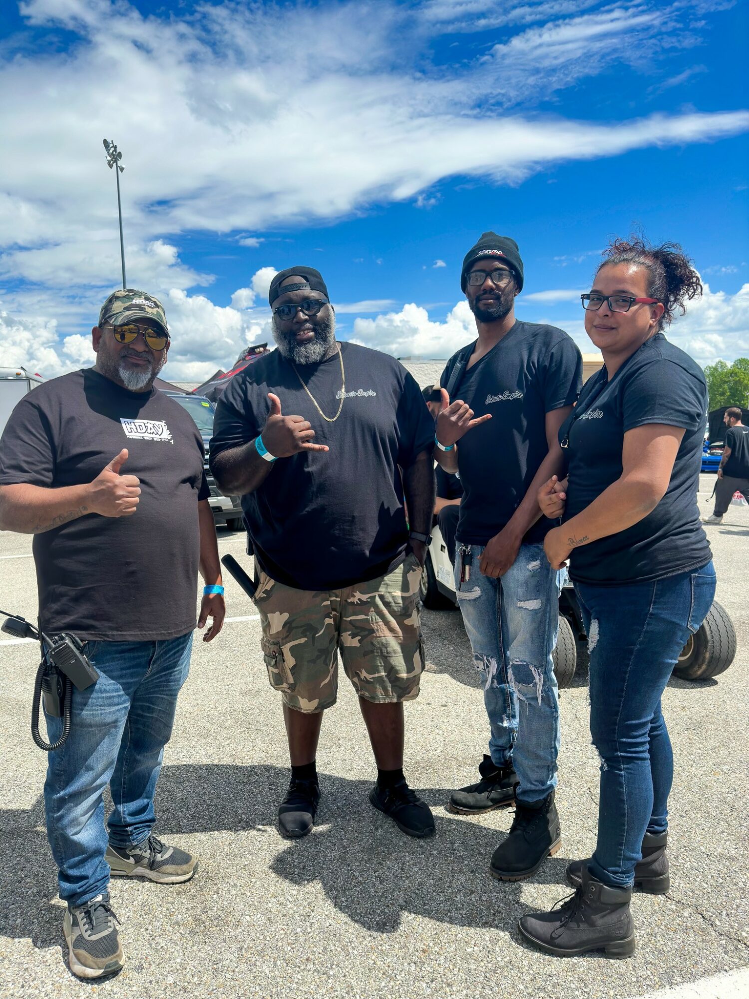 Four people standing outdoors in a parking lot under a partly cloudy sky, posing for a photo, some giving thumbs up, casual clothing.