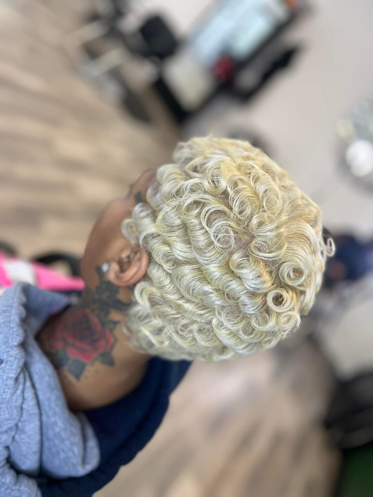 Person with short, tightly curled platinum blonde hair, holding their head with a tattooed arm visible.