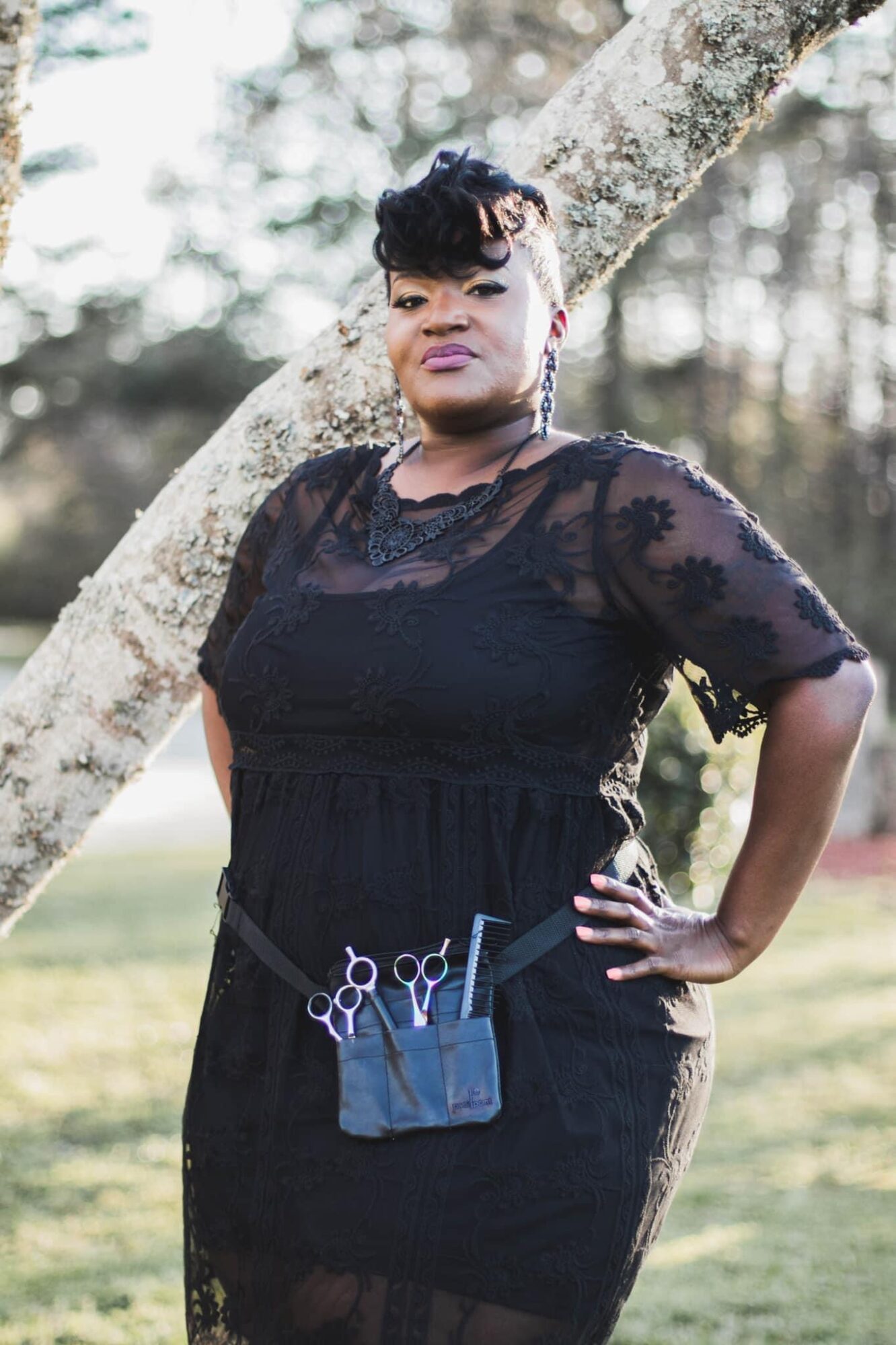 Woman in black lace dress standing outdoors near a tree with hands on hips.