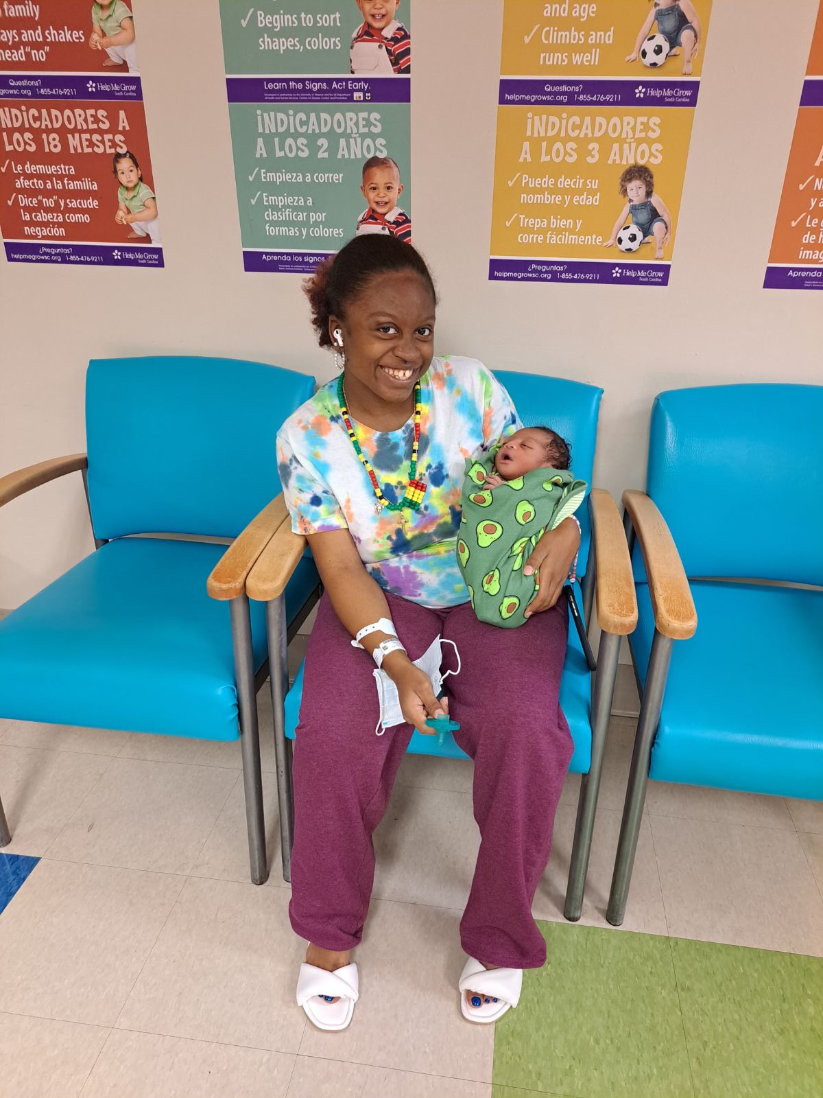 Woman sitting on a blue chair holding a baby wrapped in a green blanket, smiling, in a waiting area with colorful posters.