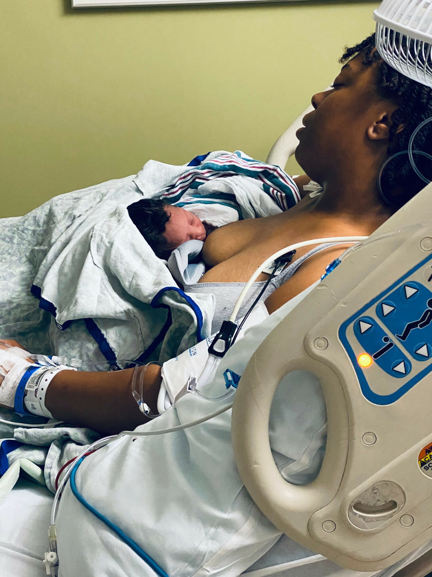 Woman in hospital bed holding newborn baby, wearing hospital gown and cap, with medical equipment nearby.