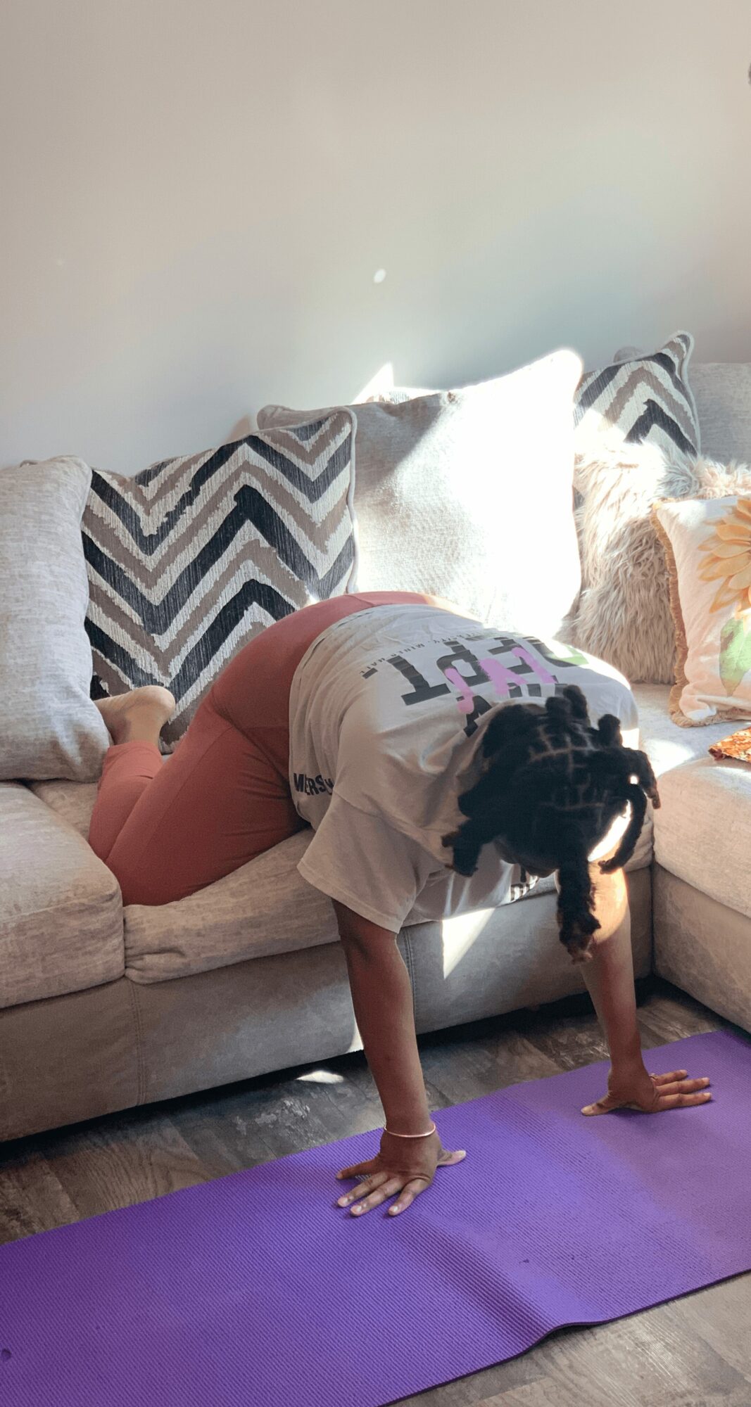 Person practicing yoga in downward dog pose on purple mat in living room, with sofa and cushions in background.