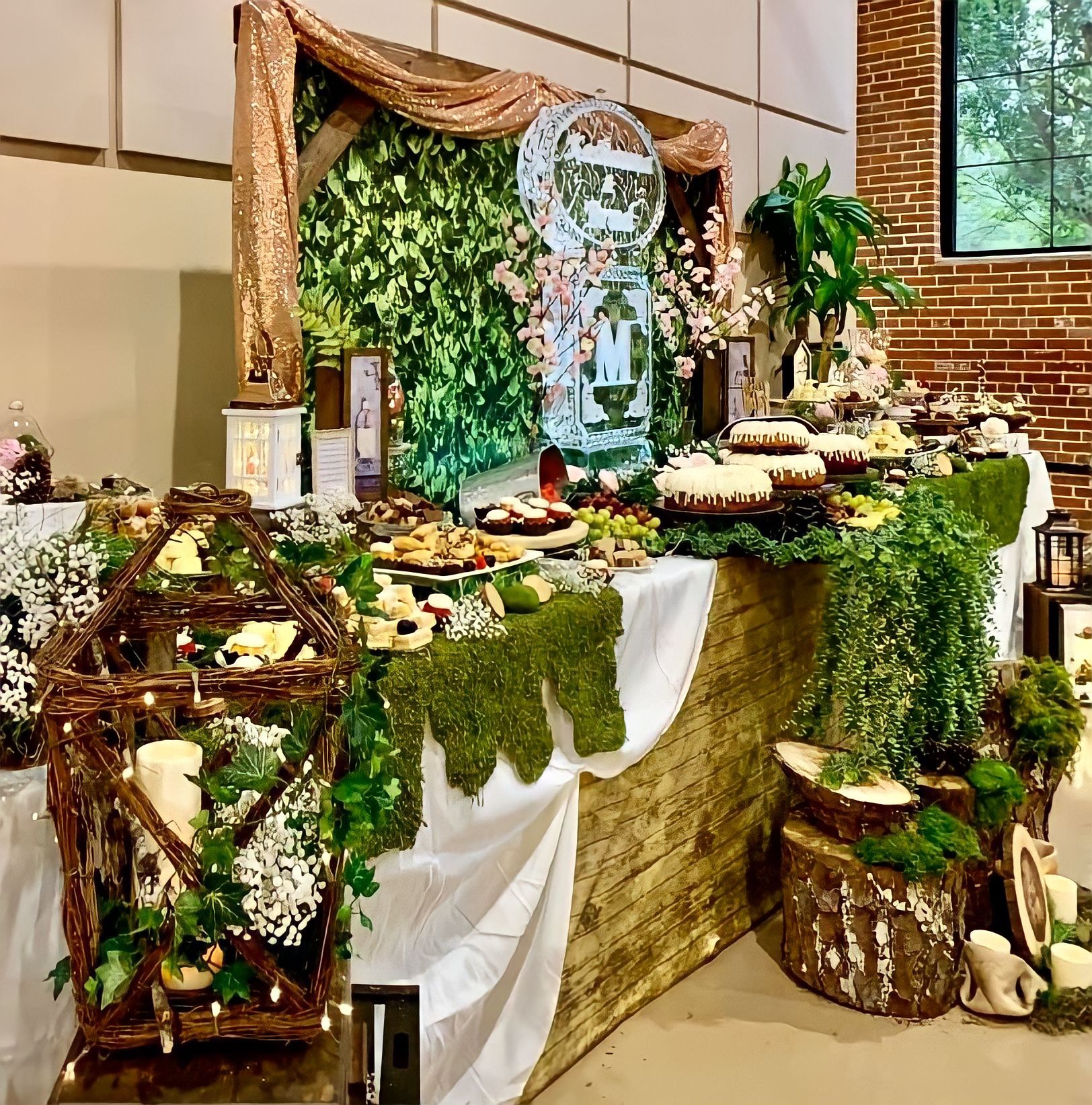 Decorative table with greenery, flowers, and desserts, set against a brick wall and window, with a rustic and elegant theme.