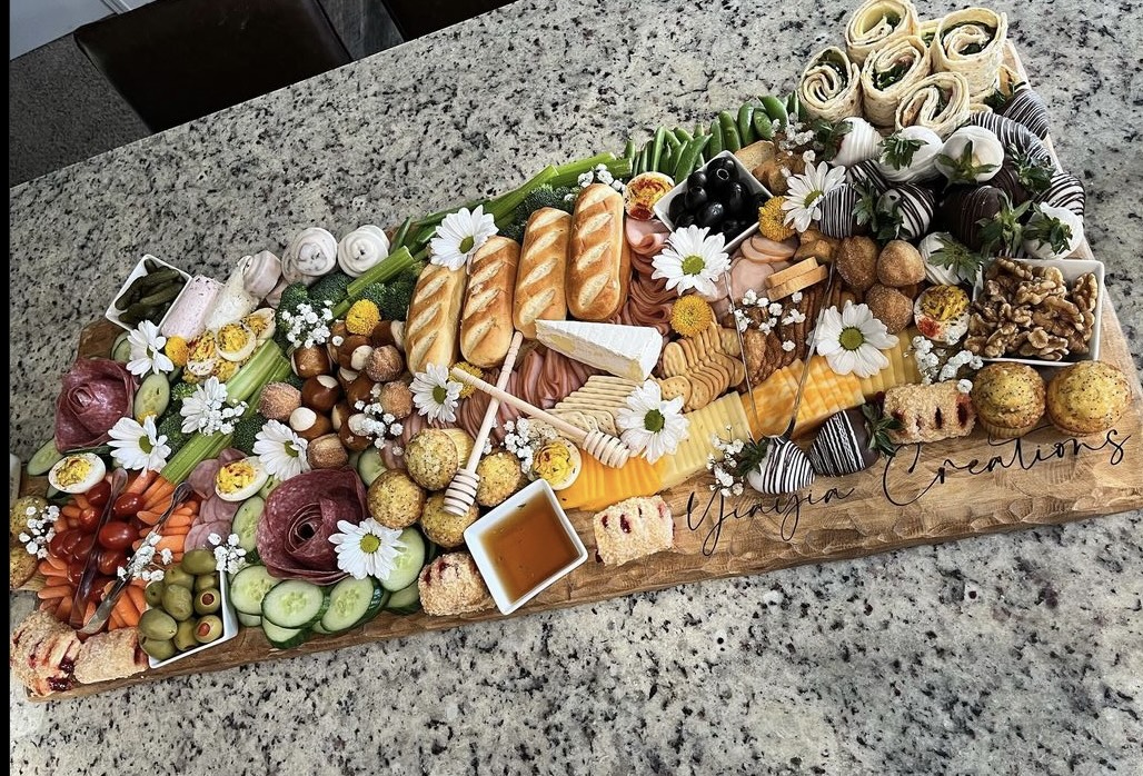 A long wooden board with assorted cheeses, fruits, nuts, and flowers on a granite countertop.