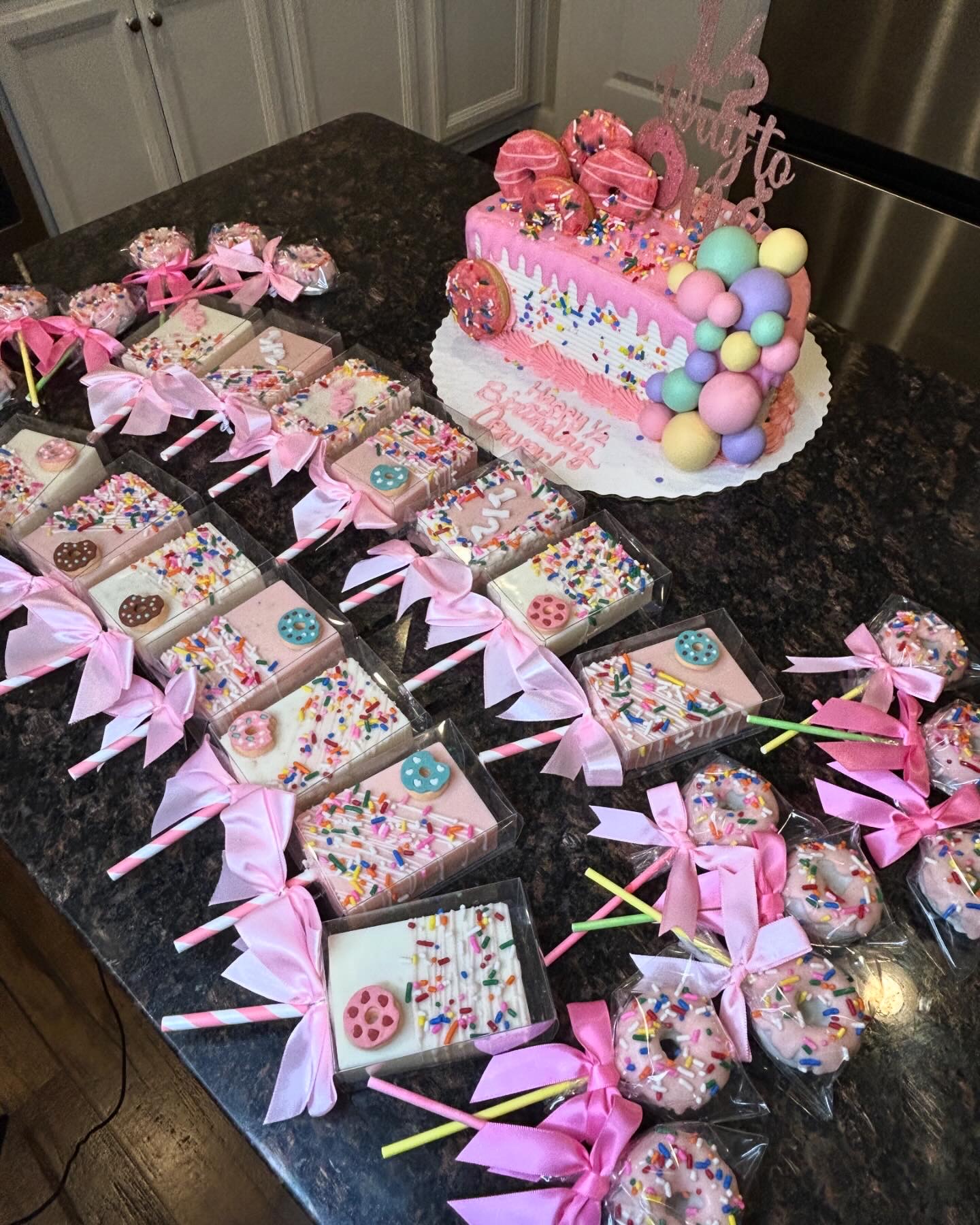 Decorated birthday cake with pink flowers and pastel balls, surrounded by wrapped gifts with pink ribbons on a black countertop.