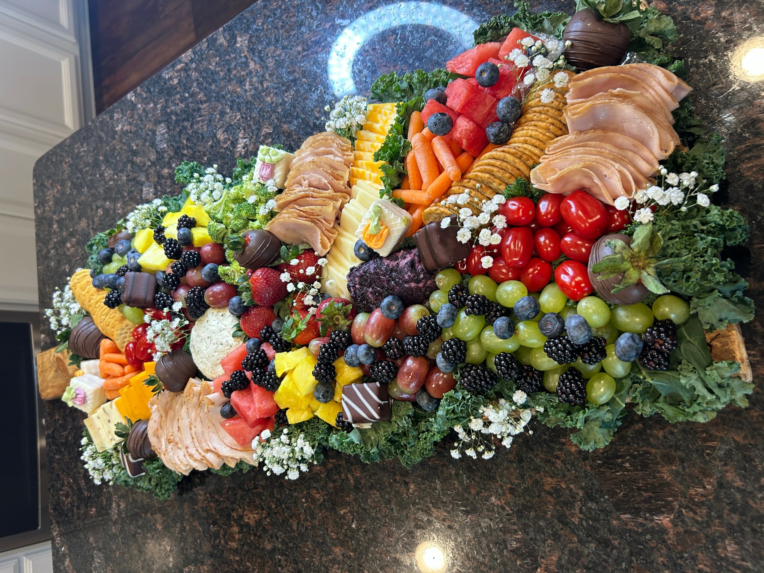 Colorful fruit and cheese platter with grapes, berries, sliced meats, and decorative garnishes on a dark surface.
