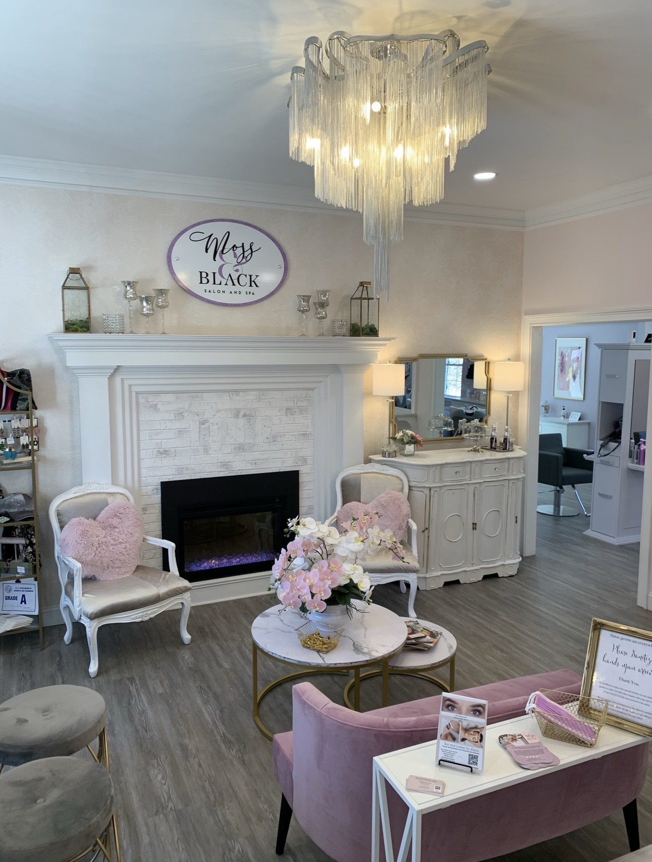 Living room with fireplace, chandelier, pink and gray chairs, floral centerpiece, and a white sideboard, decorated in soft pastel tones.