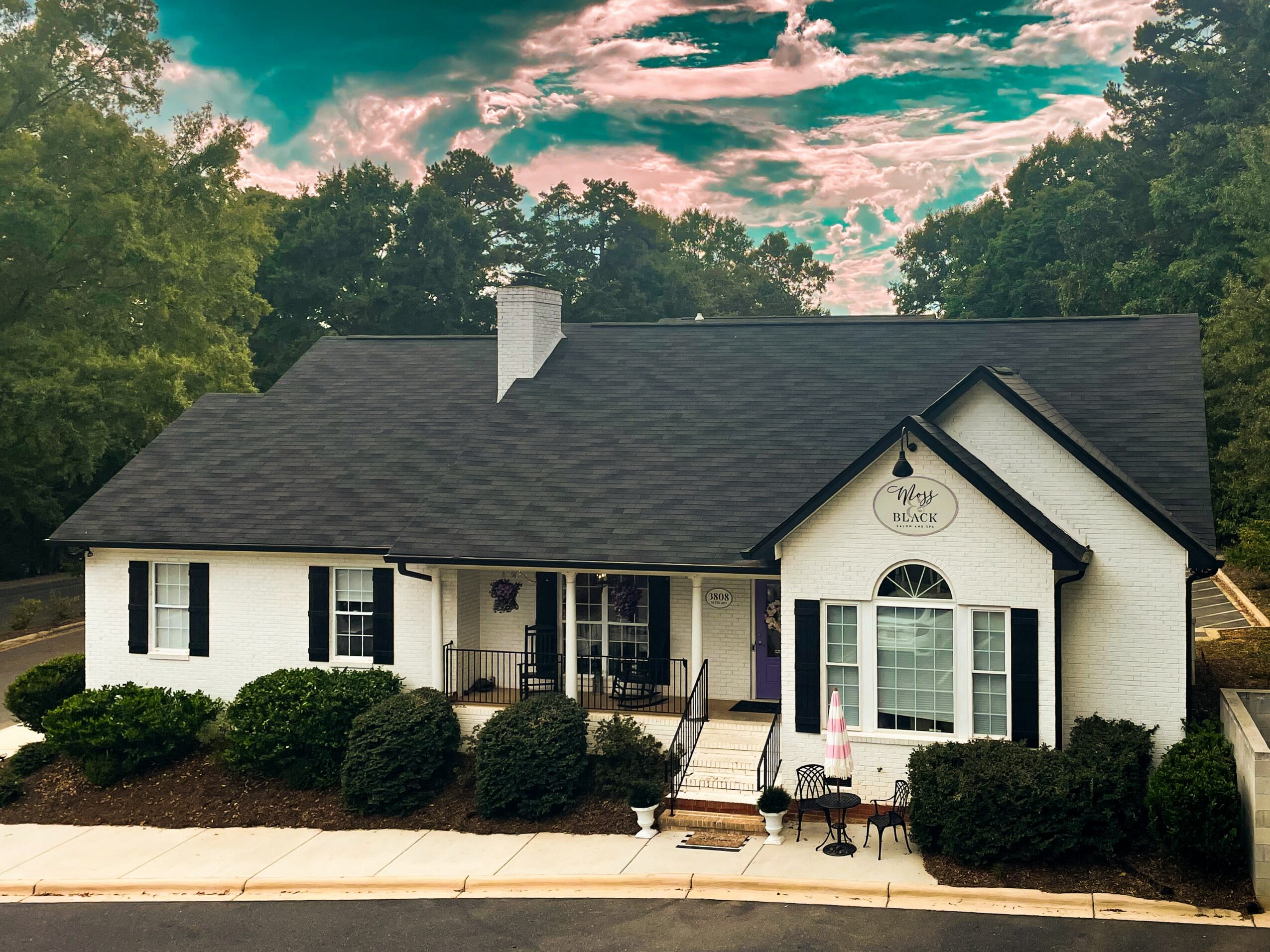 Single-story house with white exterior, black roof, front porch, and landscaped bushes, under a partly cloudy sky.