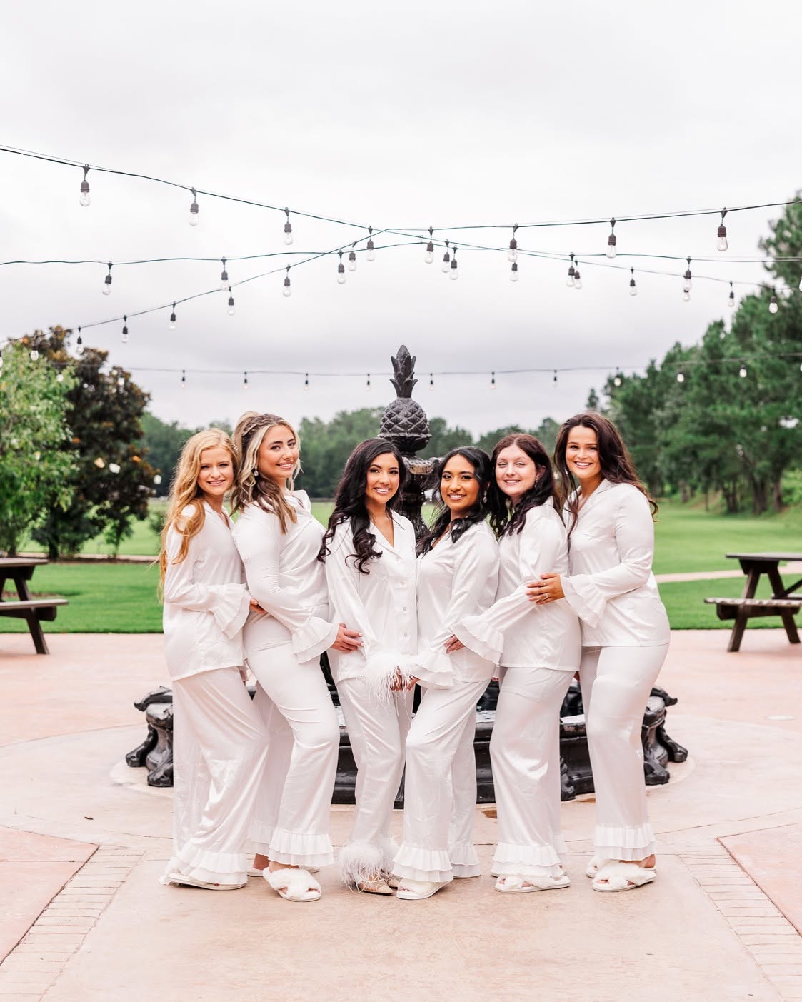 Six women in white outfits standing outdoors on a paved area with trees and string lights overhead.