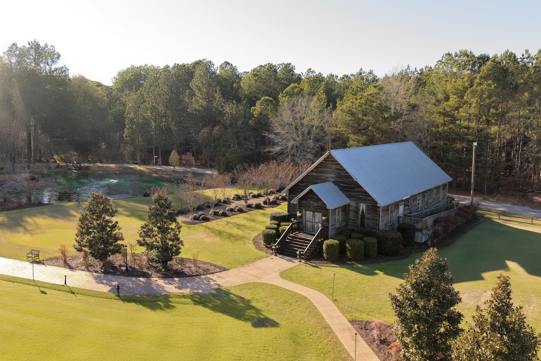 A stone building with a metal roof is surrounded by a park with trees, pathways, and a grassy area.
