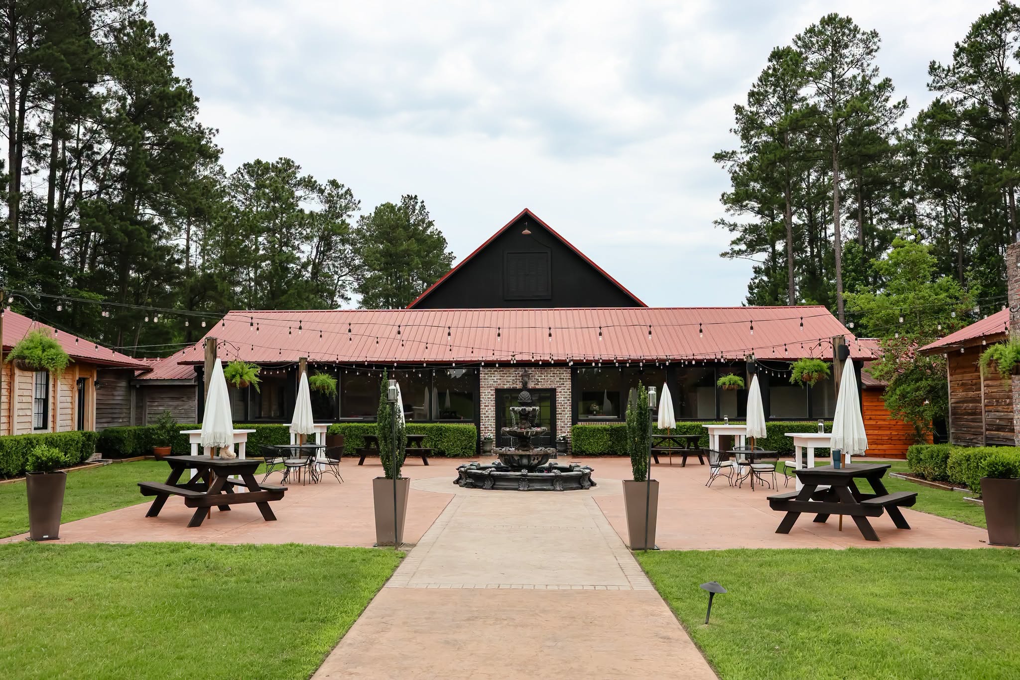 Pathway leading to a building with a red roof, surrounded by picnic tables, umbrellas, and trees, under a cloudy sky.
