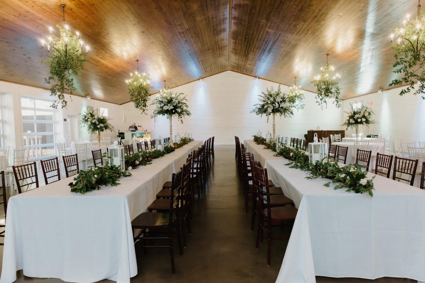 Long banquet tables with white tablecloths, decorated with greenery and floral arrangements, in a warmly lit room with wooden ceiling and chandeliers.