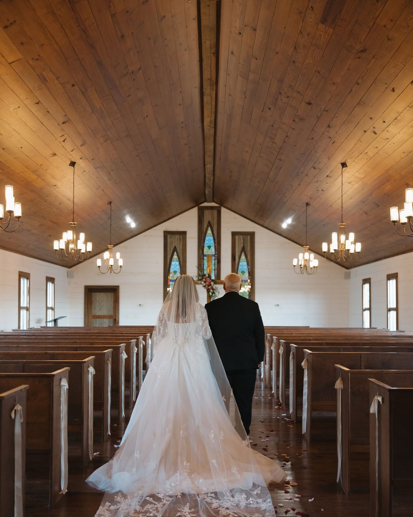 Bride and groom walking down the aisle in a church with wooden ceiling and pews, stained glass windows, and chandeliers.