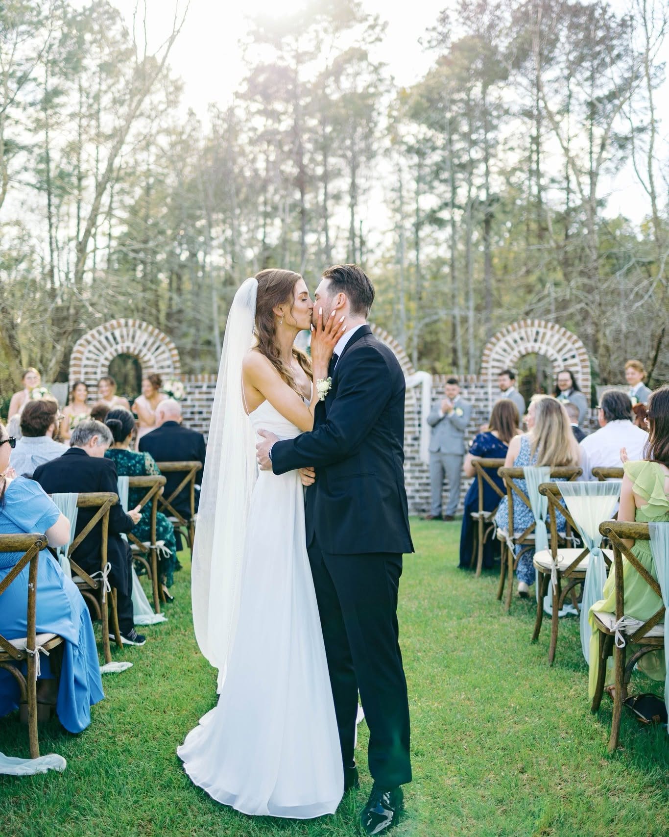Bride and groom kiss outdoors during wedding ceremony, guests seated on chairs, trees in background.