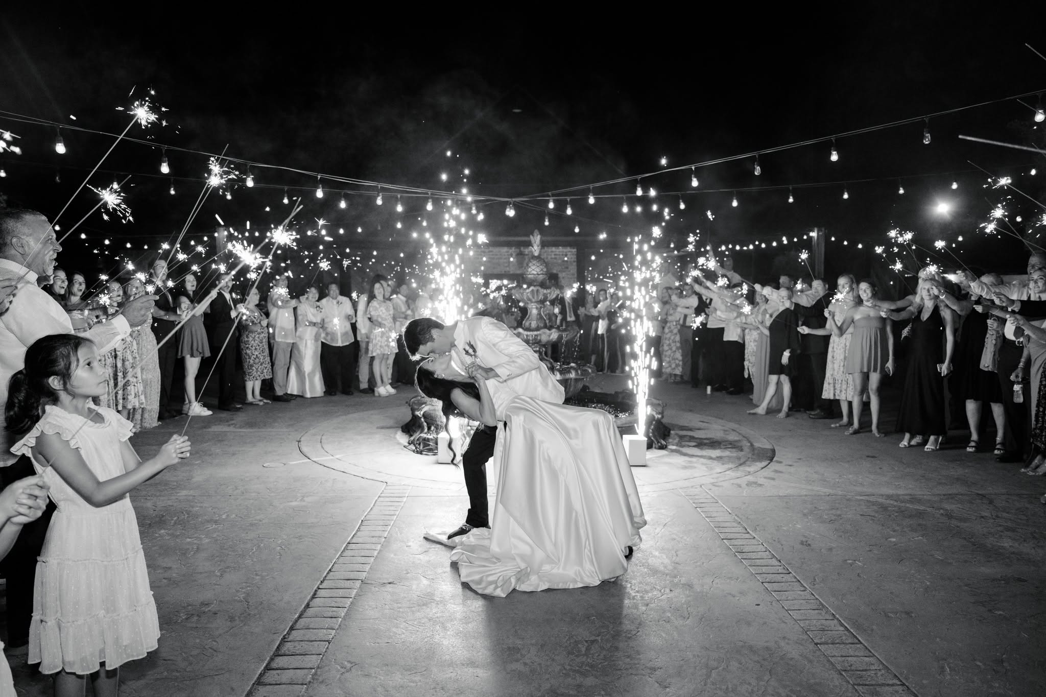 Couple dancing surrounded by people, with sparklers and string lights overhead, in a nighttime celebration, black and white photo.
