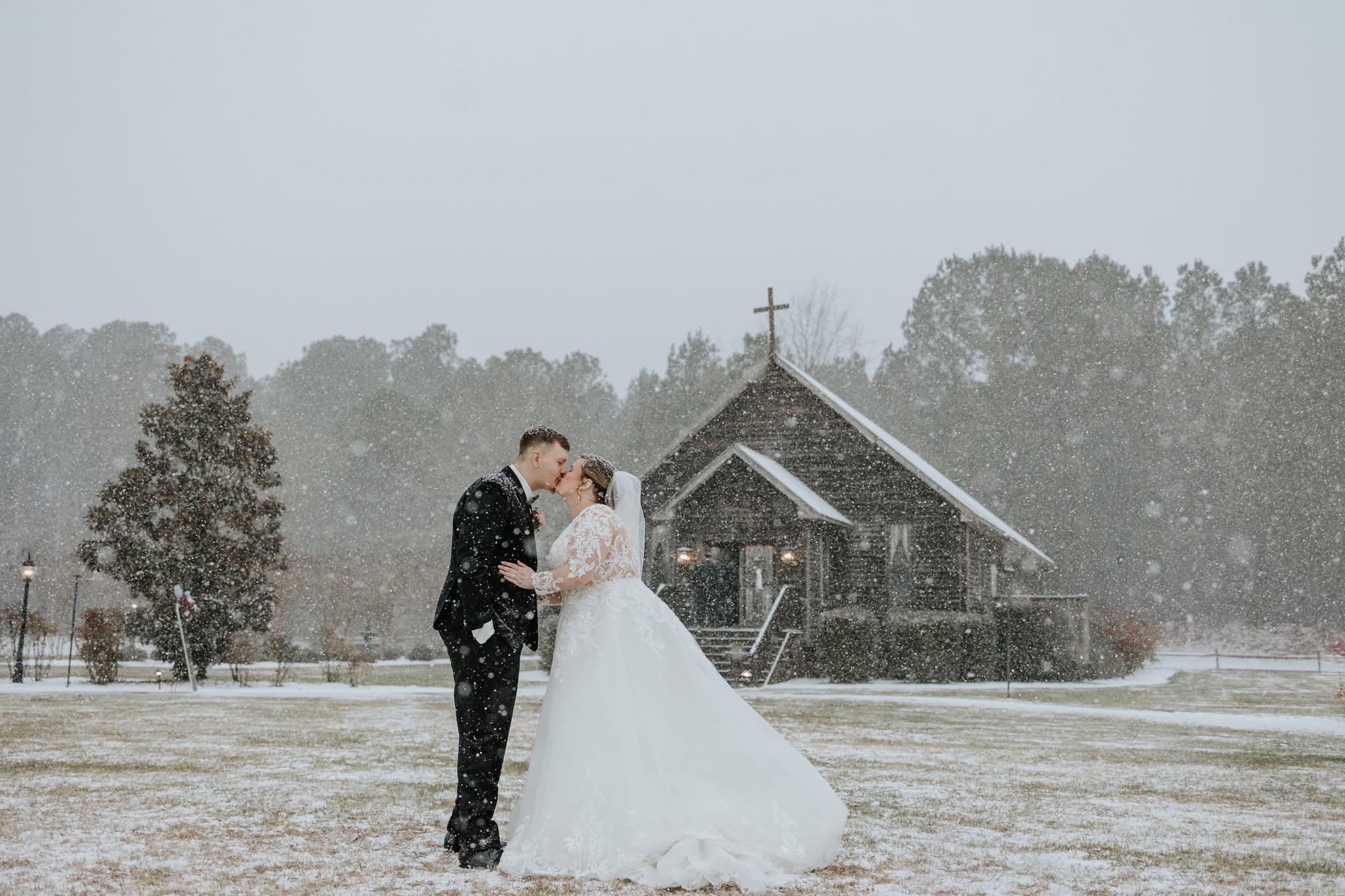 Bride and groom kiss outdoors in snow, church in background, trees, overcast sky, winter scene, wedding attire, snow falling