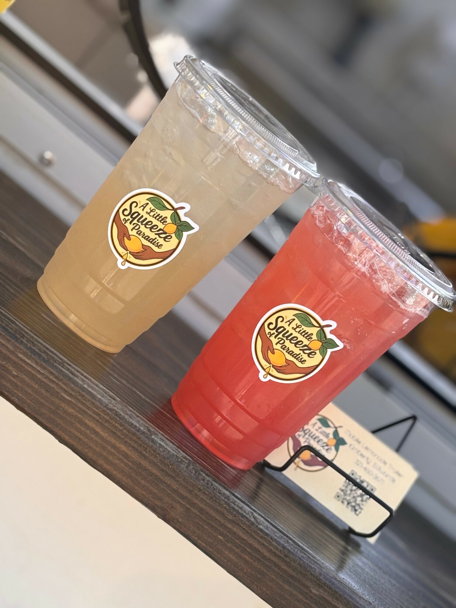 Two plastic cups with fruit juice drinks on a table, one yellow and one pink, with a logo sticker, a card, and a blurred background.