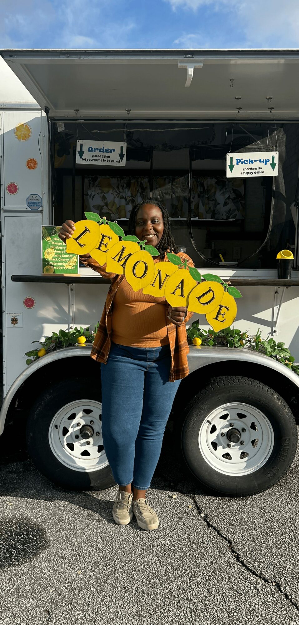 Woman standing in front of a lemonade stand holding a yellow sign that says 'LEMONADE'.
