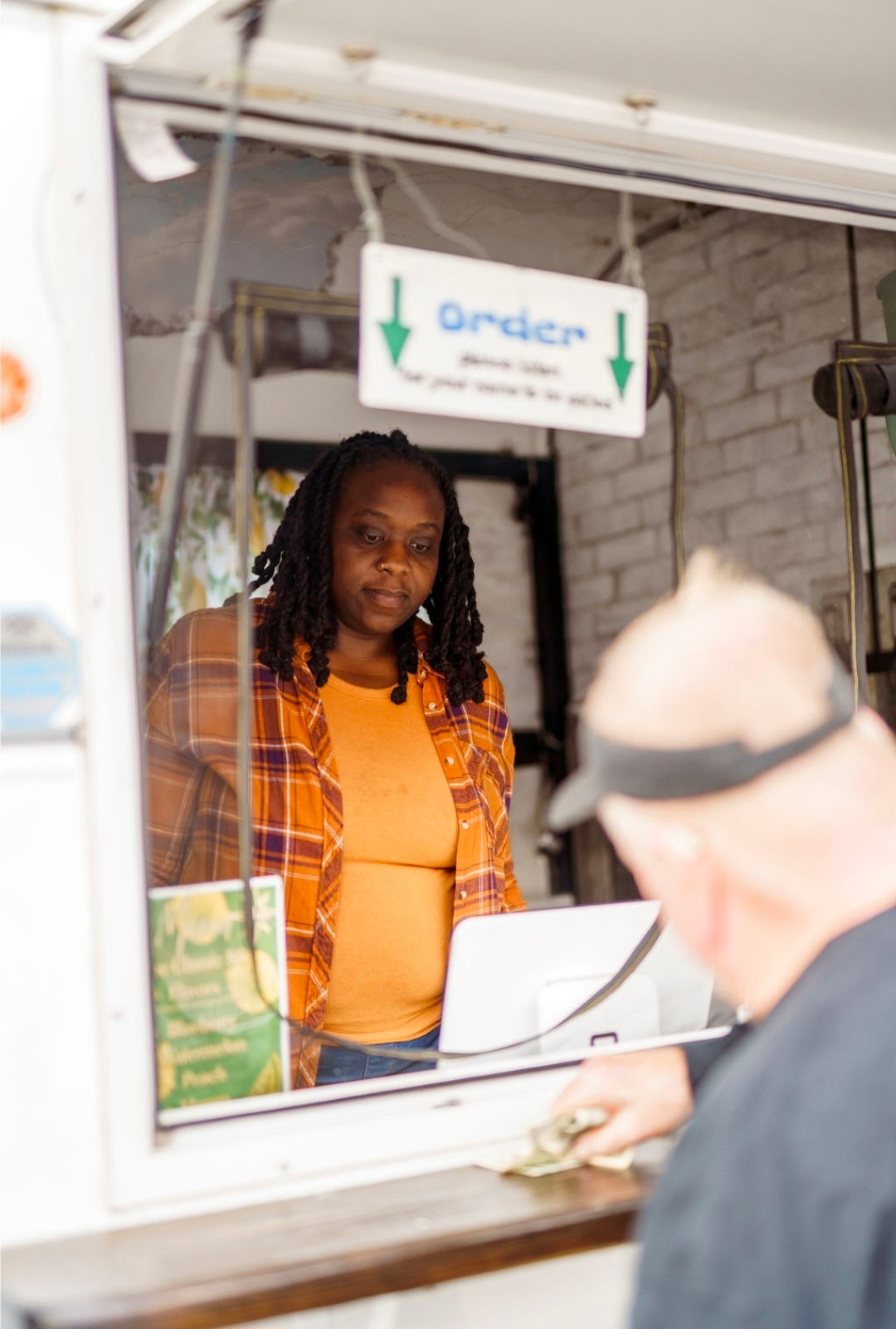 Woman with long braids wearing orange plaid shirt and yellow top, standing behind a counter, looking at a person in front.