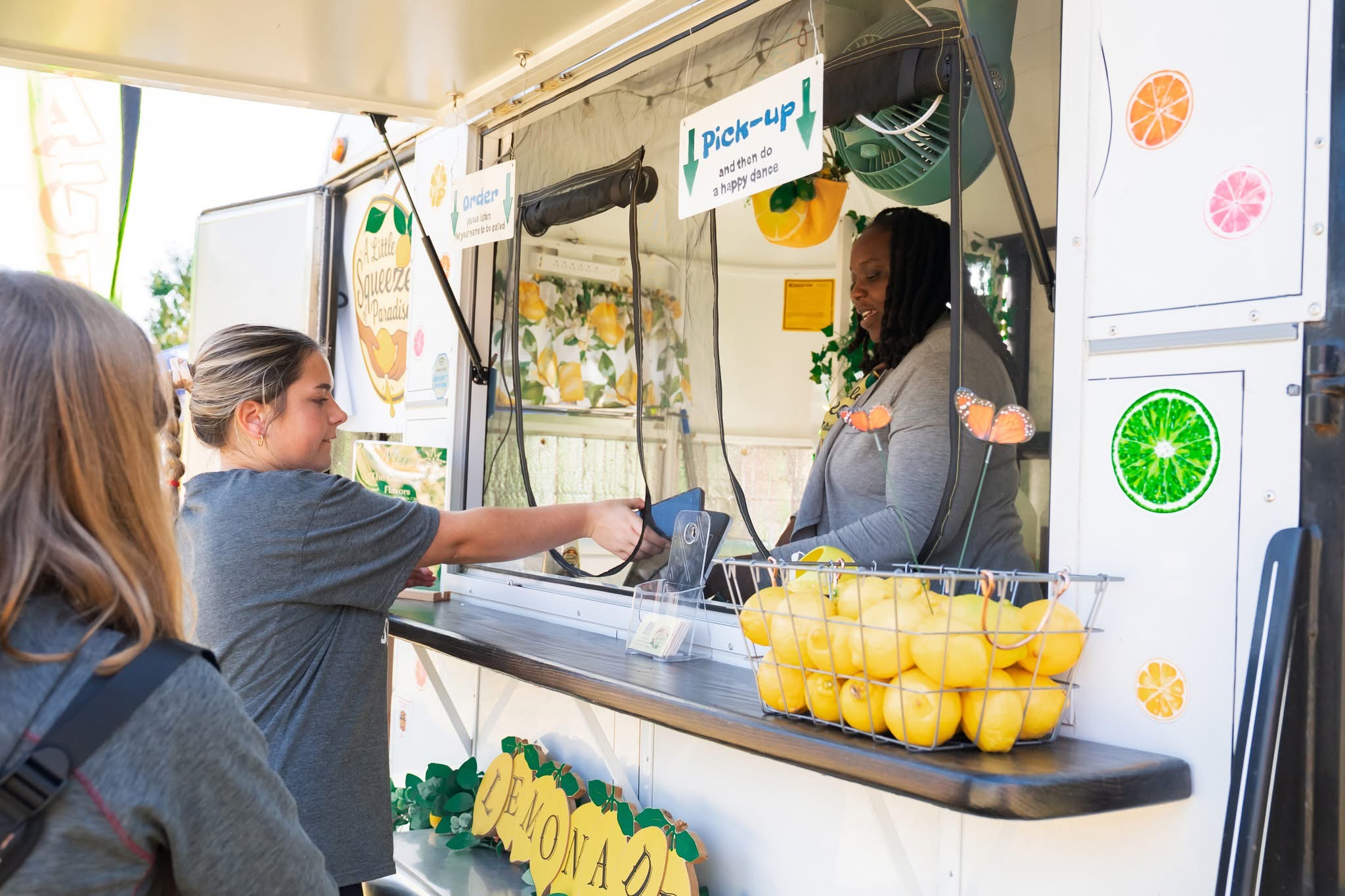 Two women at a lemon stand exchanging money, with lemons in a bowl and colorful decorations around.