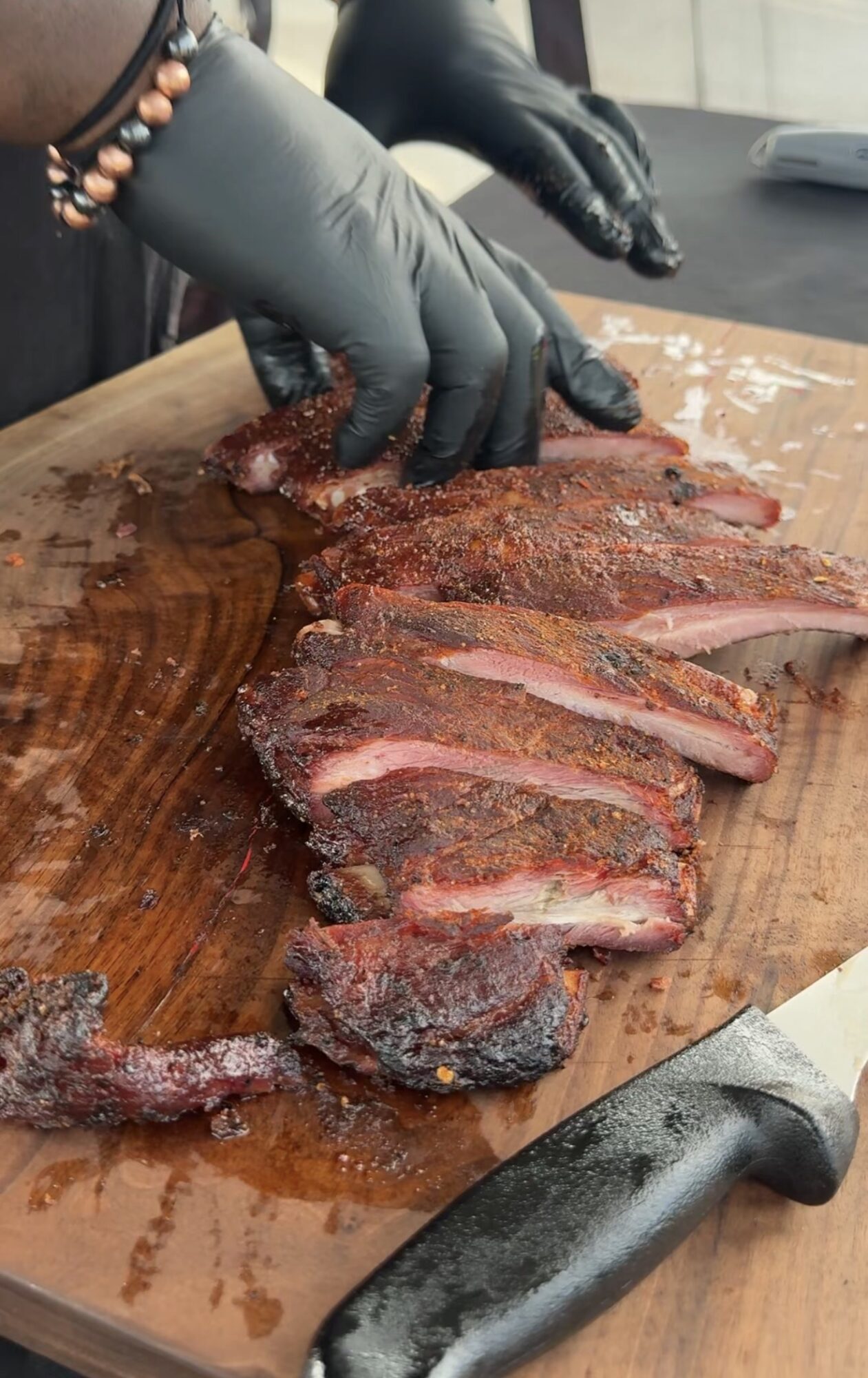 Person slicing cooked meat on a wooden cutting board, wearing black gloves and a beaded bracelet.
