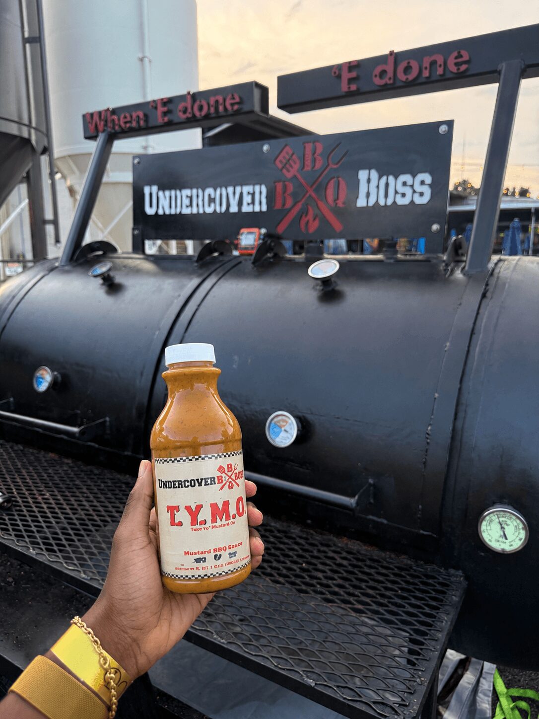 Hand holding a bottle of T.Y.M. juice in front of a black barbecue smoker with signs reading 'When E done' and 'E done'.