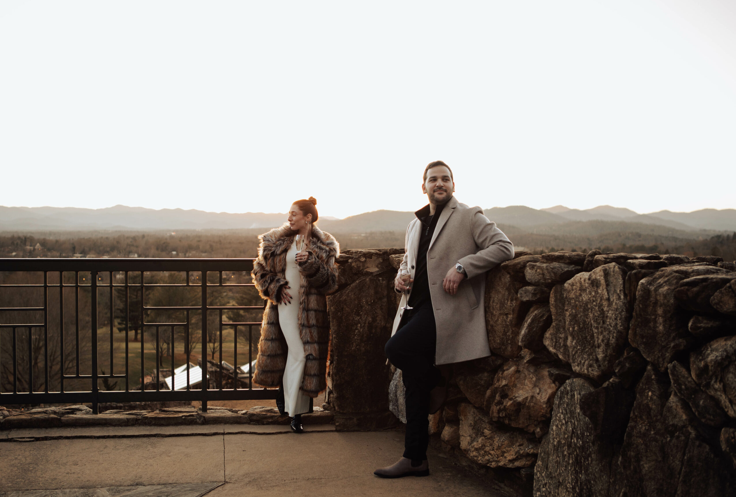 Two people standing outdoors near a stone wall and railing, with a landscape and mountains in the background.
