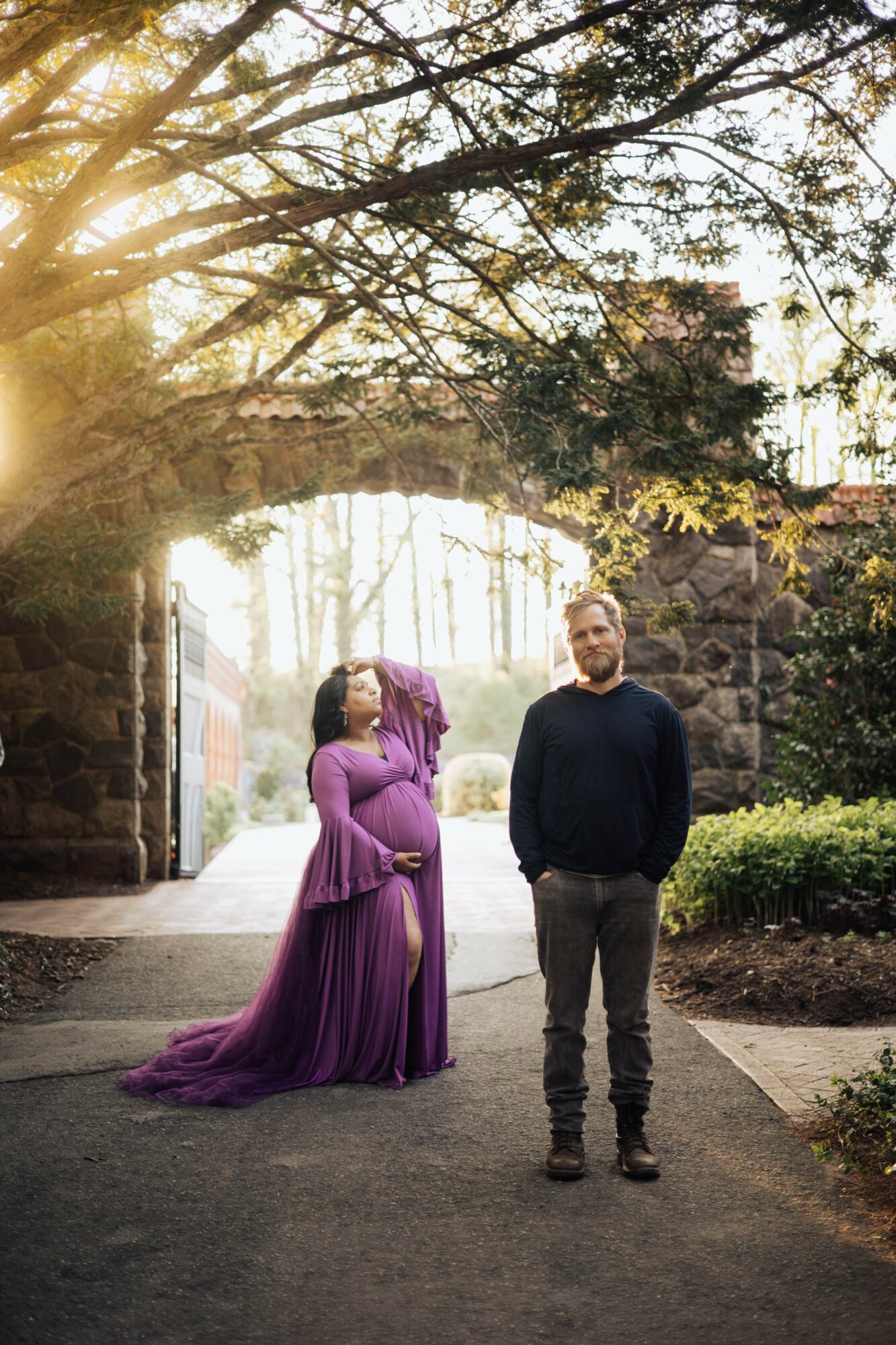 Two people standing outdoors under a tree archway, one in a purple dress, the other in a dark sweater and pants, sunlight filtering through leaves.