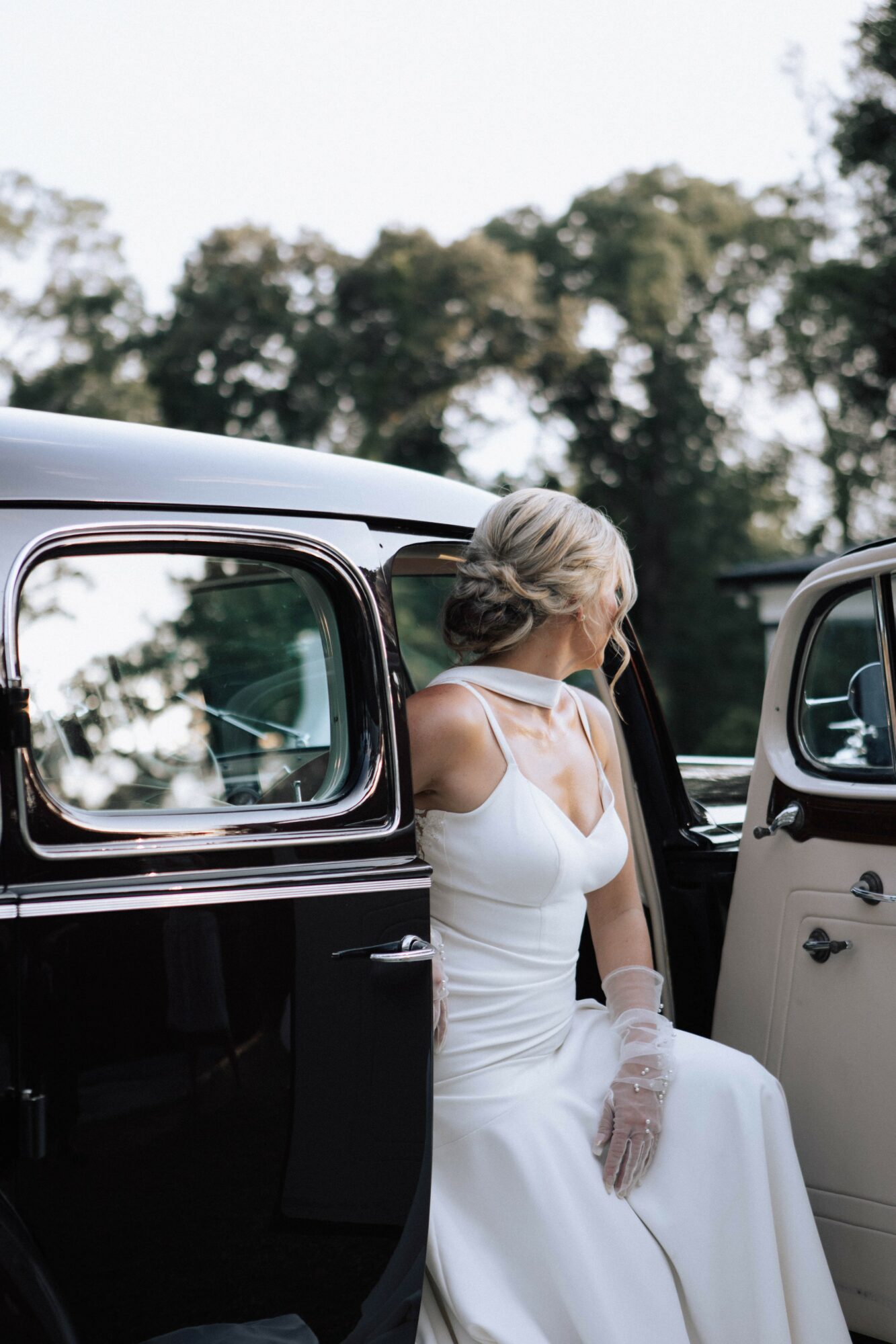 Woman in white dress and gloves standing outside vintage cars, trees in background.