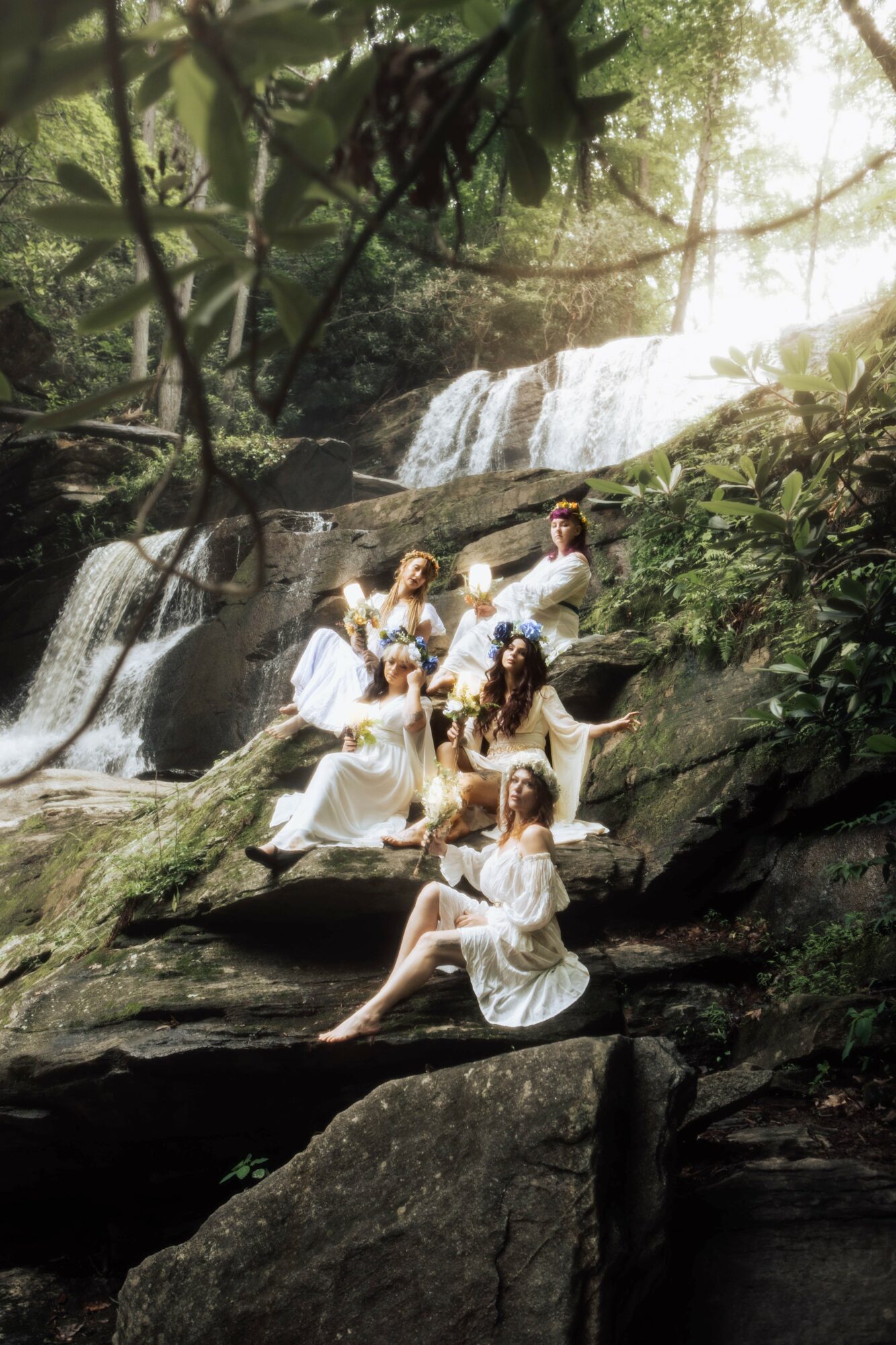 Group of women in white dresses sitting on rocks near a waterfall in a forest, sunlight filtering through trees.