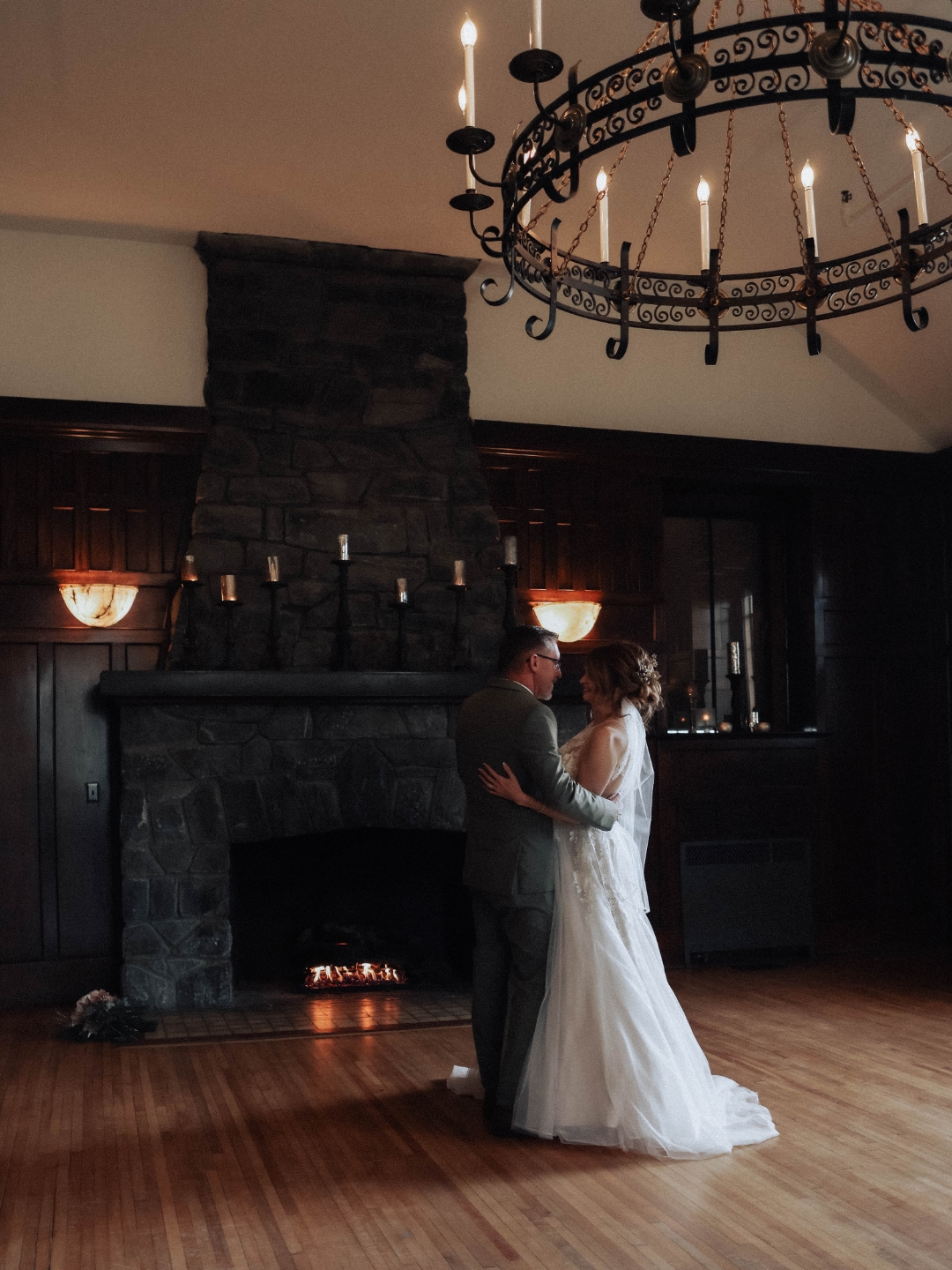 A couple in wedding attire dancing in front of a fireplace with candles, chandelier overhead, and wooden floor.