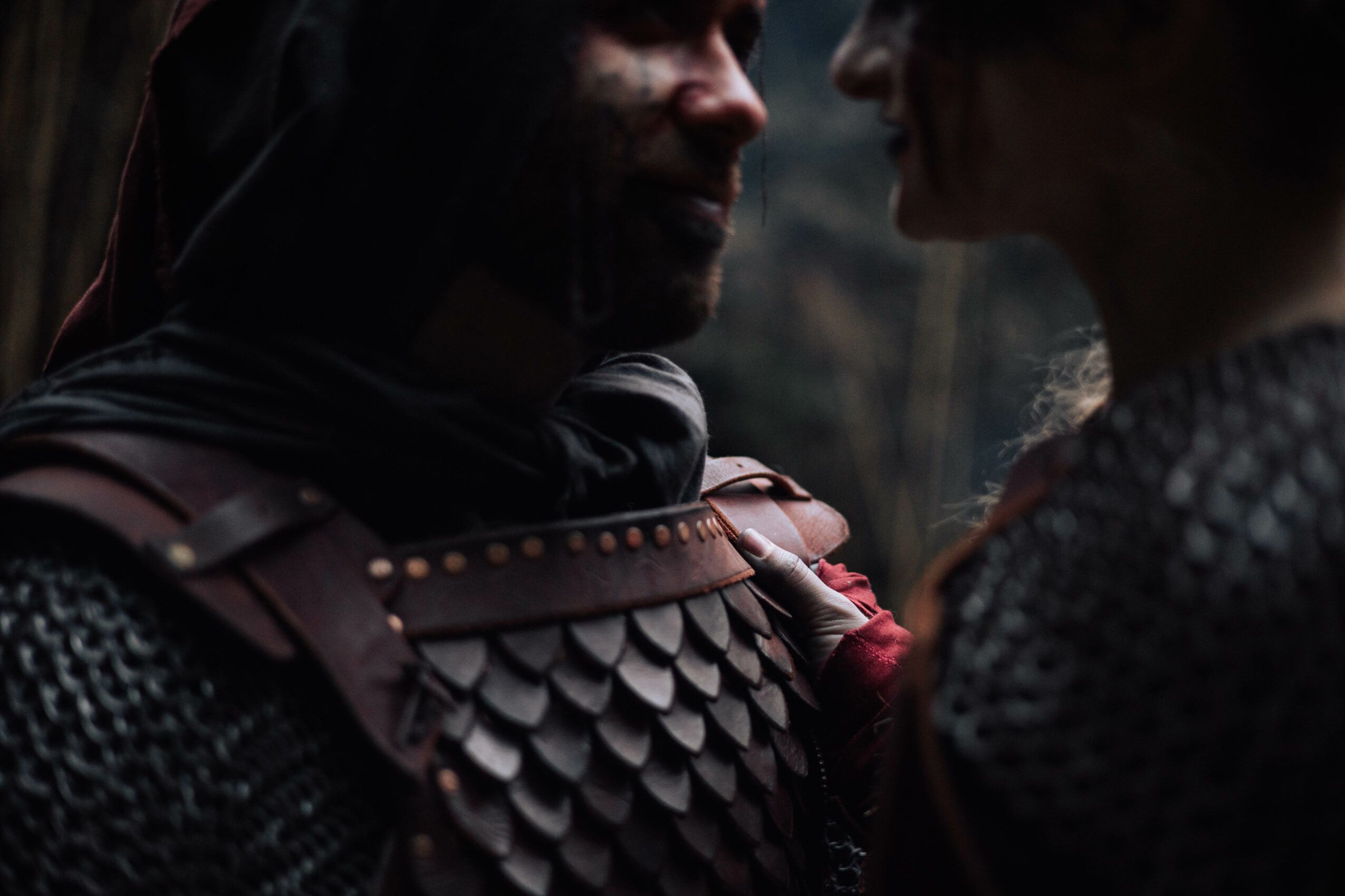 Two people face each other closely, with dark clothing and armor-like textures, in a dim outdoor setting.