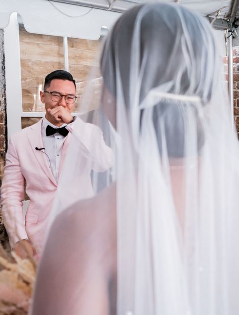Person in wedding dress and veil facing a man in a pink suit and glasses, smiling and pointing.