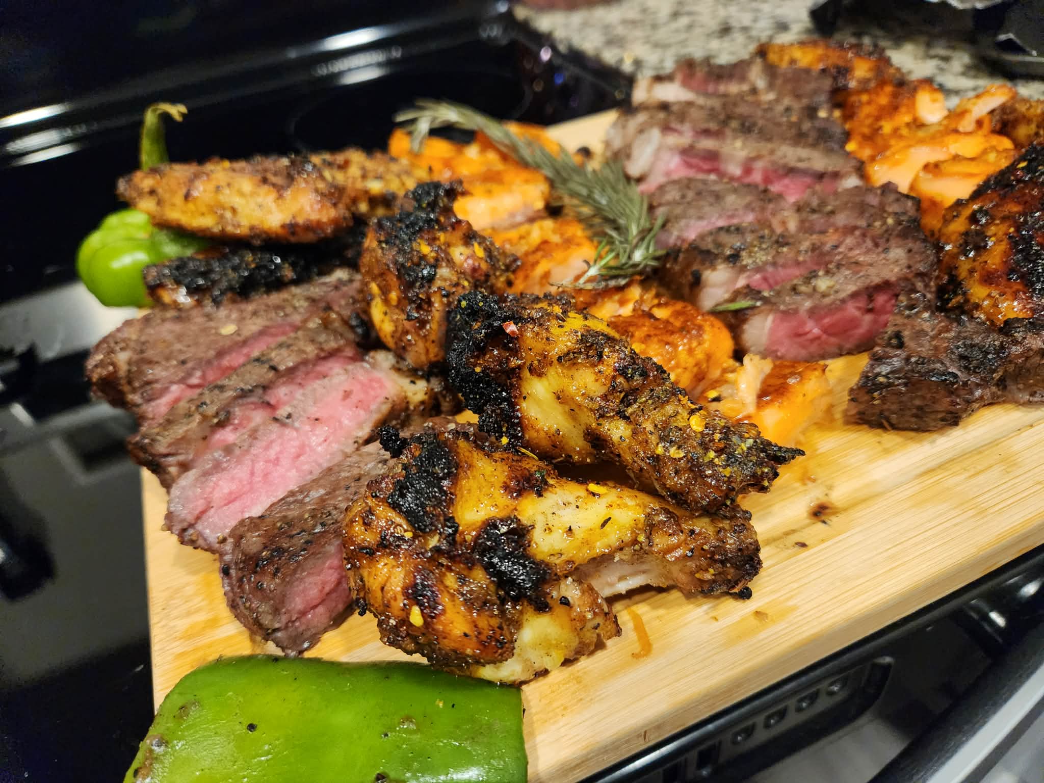 Slices of grilled steak and chicken on a wooden cutting board with green peppers, some charred, and a black tray in the background.