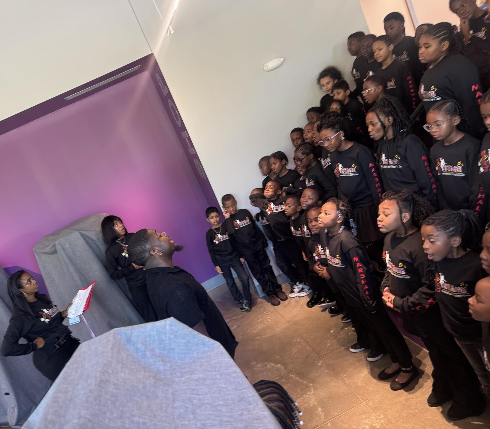 Group of children and adults gathered indoors, some children singing or reciting, adults observing, purple wall in background.