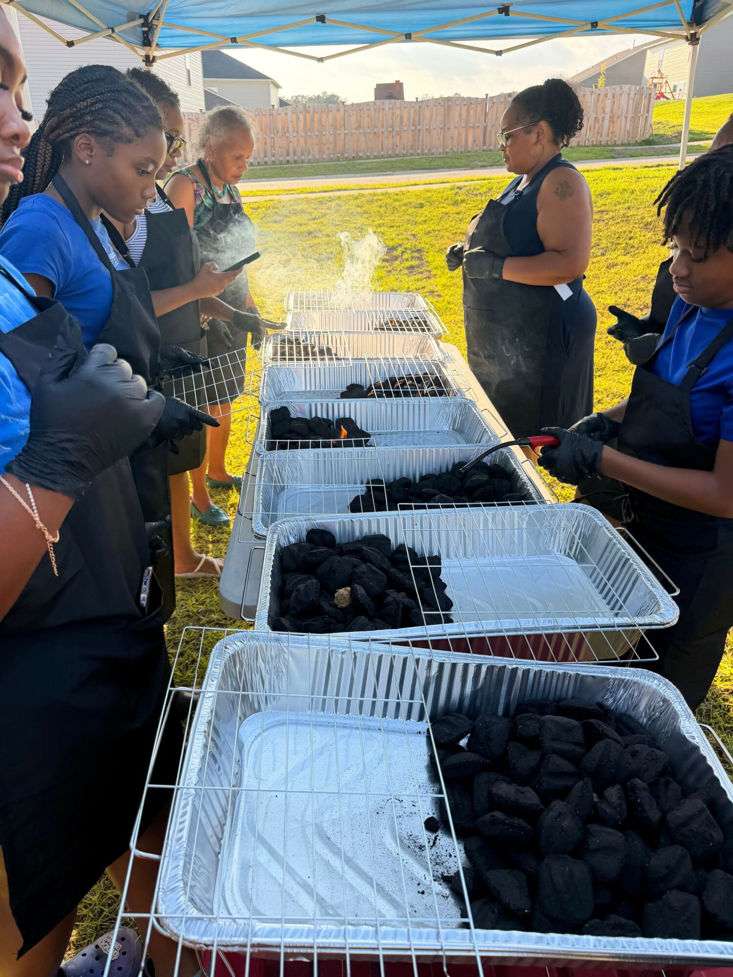 People gather outdoors around trays of black charcoal, some wearing gloves and masks, preparing to handle the charcoal.