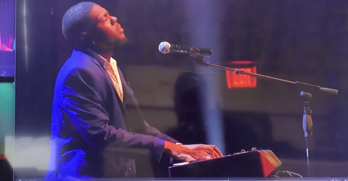 Person playing keyboard and singing into microphone on stage, with purple and blue lighting