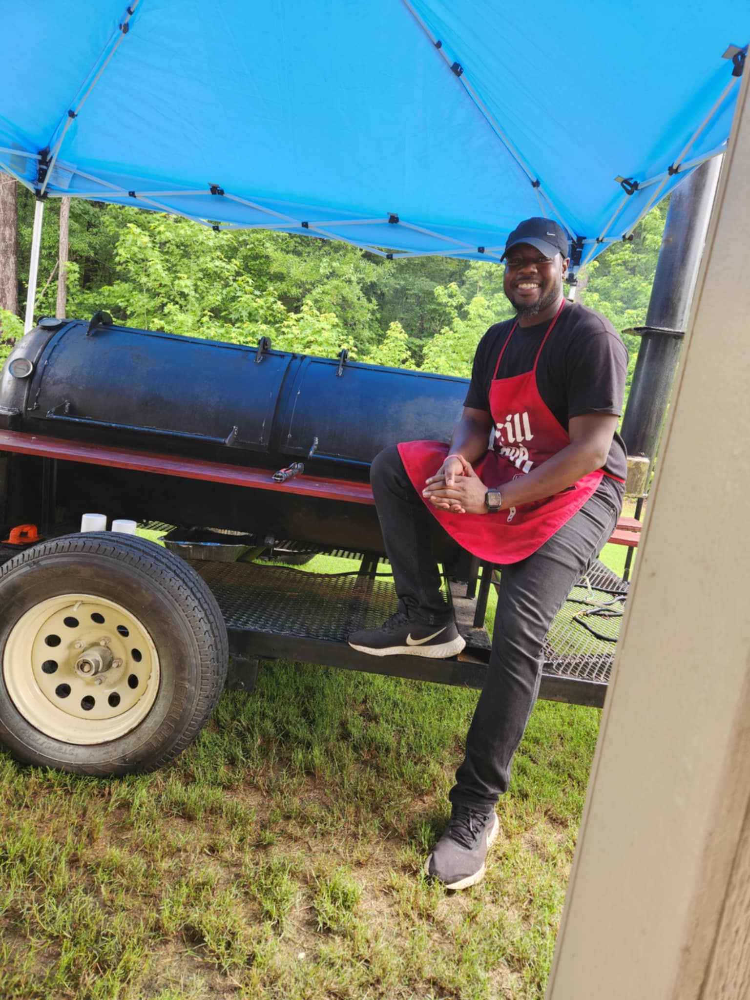 Person smiling, wearing a red apron, standing next to a large black barbecue grill under a blue canopy, outdoors.