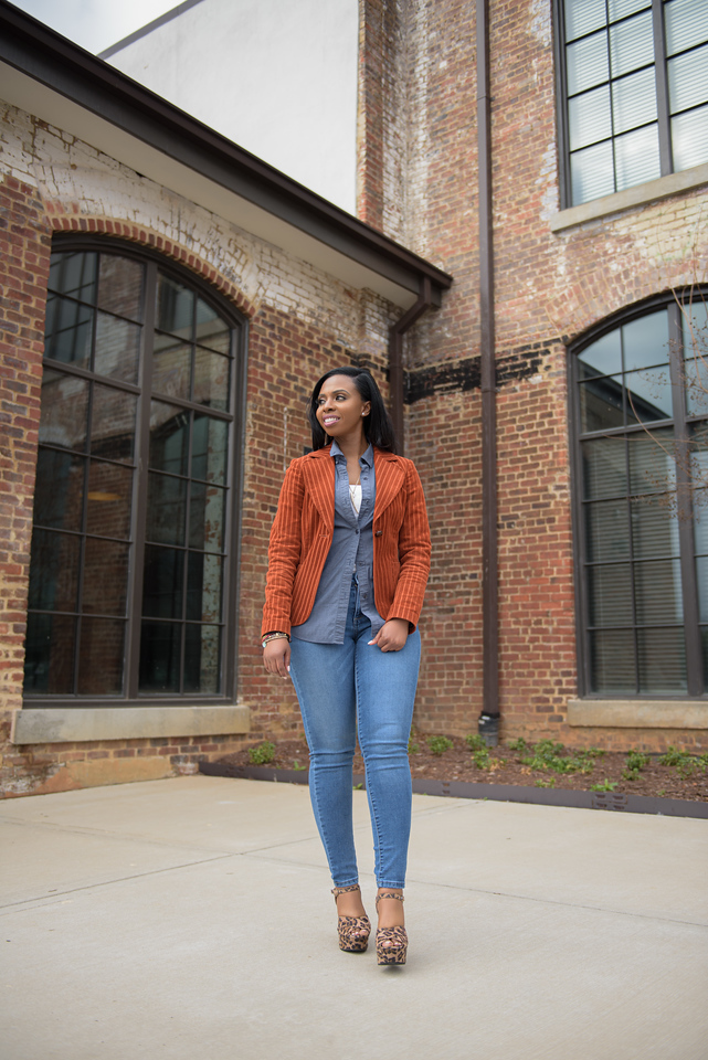 Woman standing outdoors in front of brick building with large windows, wearing a brown jacket, blue jeans, and heels.