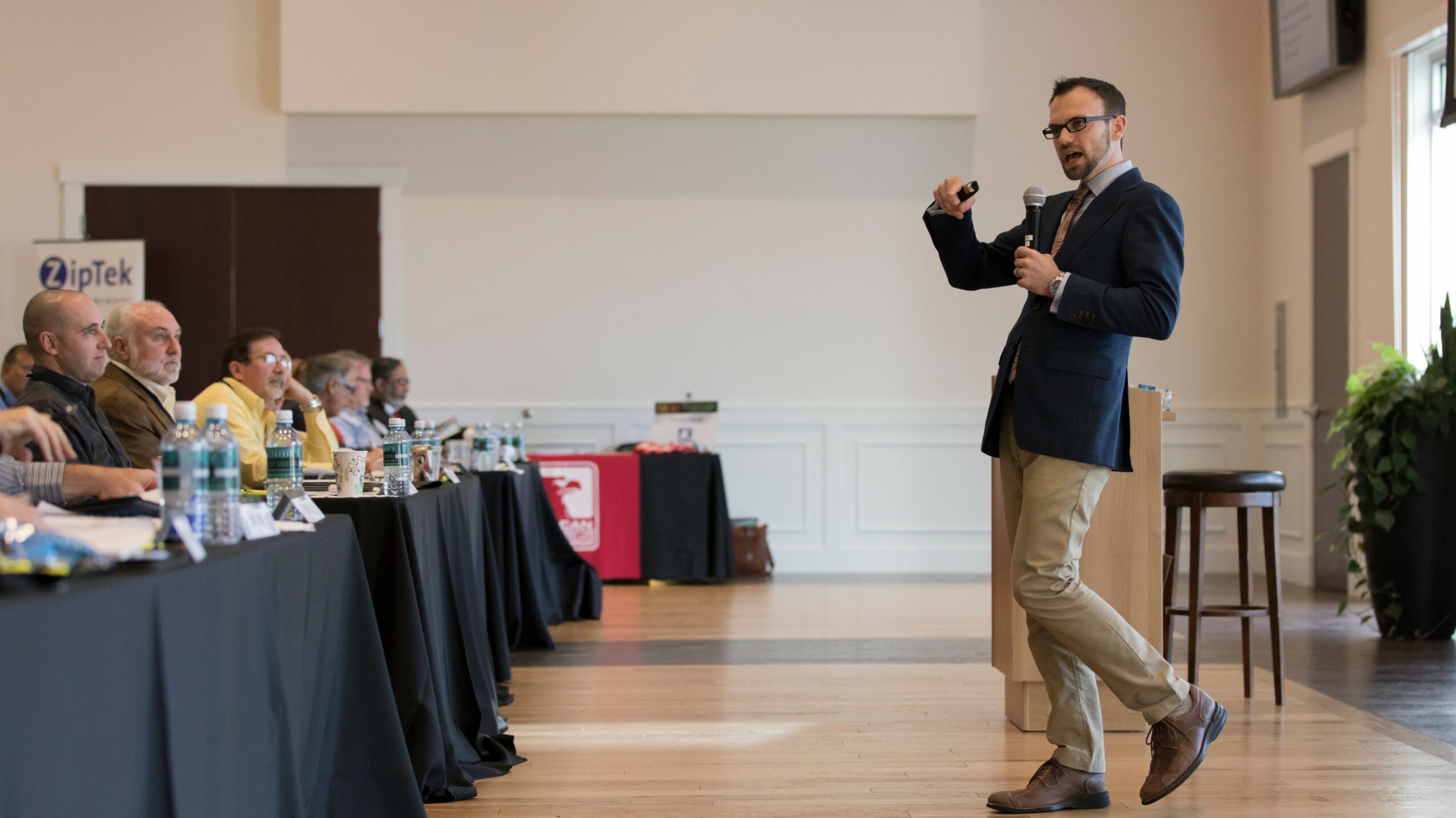 A man in a blazer and khakis stands in front of a seated audience in a conference room, gesturing with his hand.