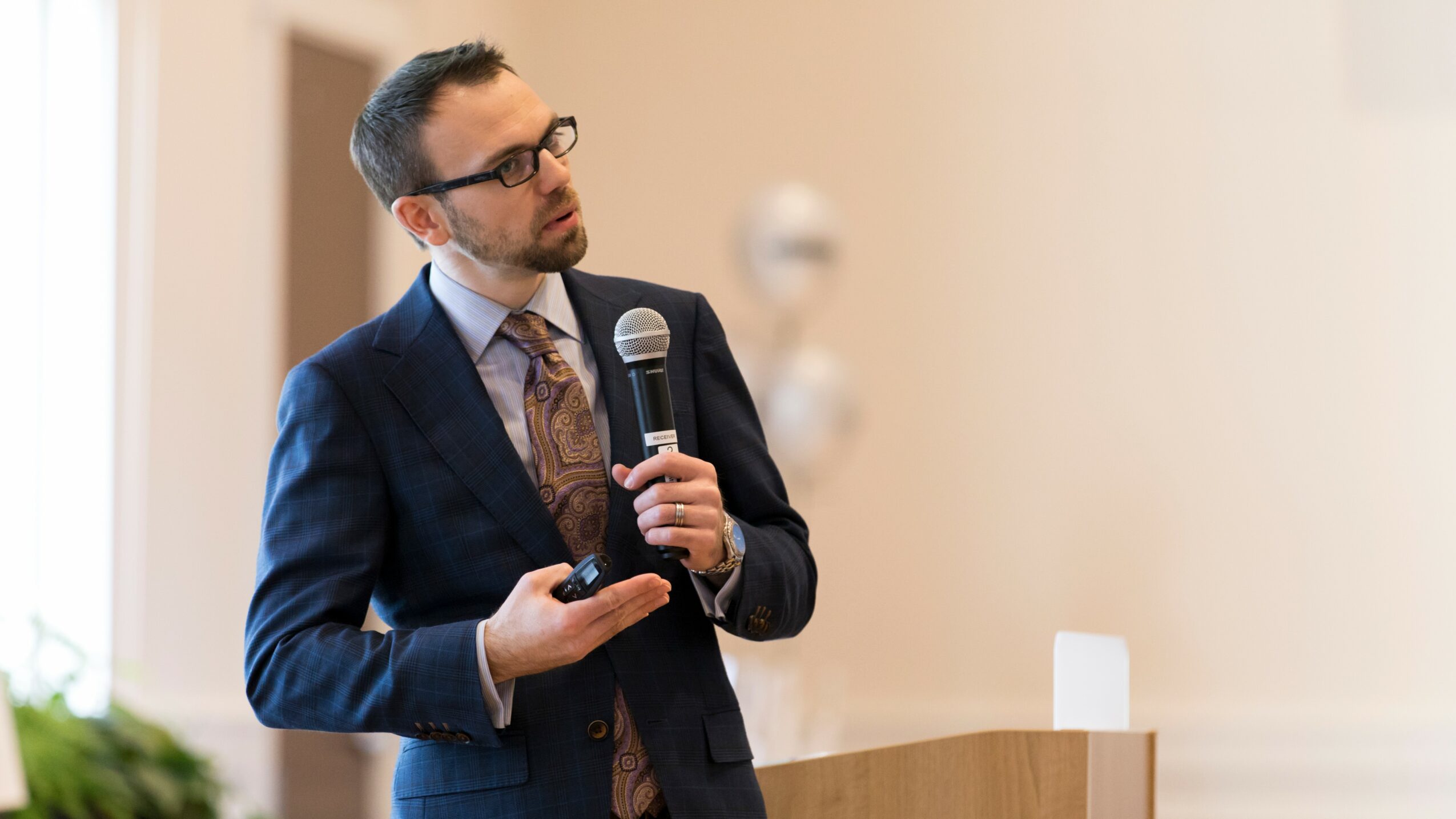 Man in suit holding microphone and remote, standing at a podium during a presentation, with a blurred background.