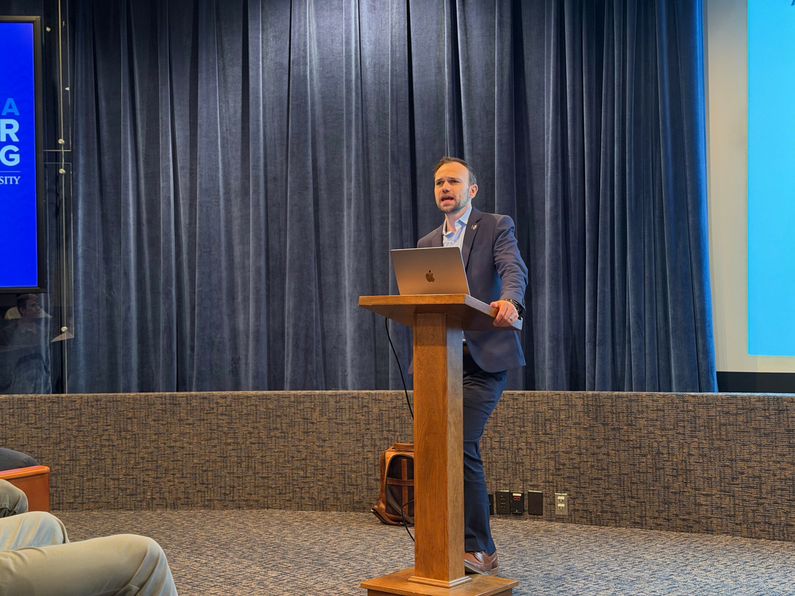Man in suit stands at podium giving a presentation in front of dark curtains, with a laptop on the podium.