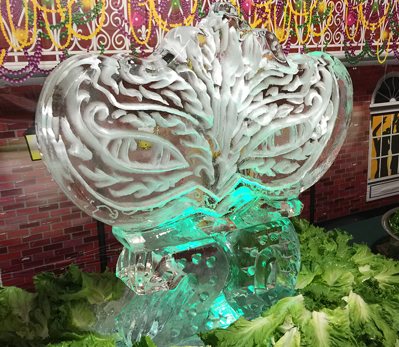 Ice sculpture of a butterfly with detailed wings, illuminated with green light, surrounded by lettuce leaves, outside a decorated building.