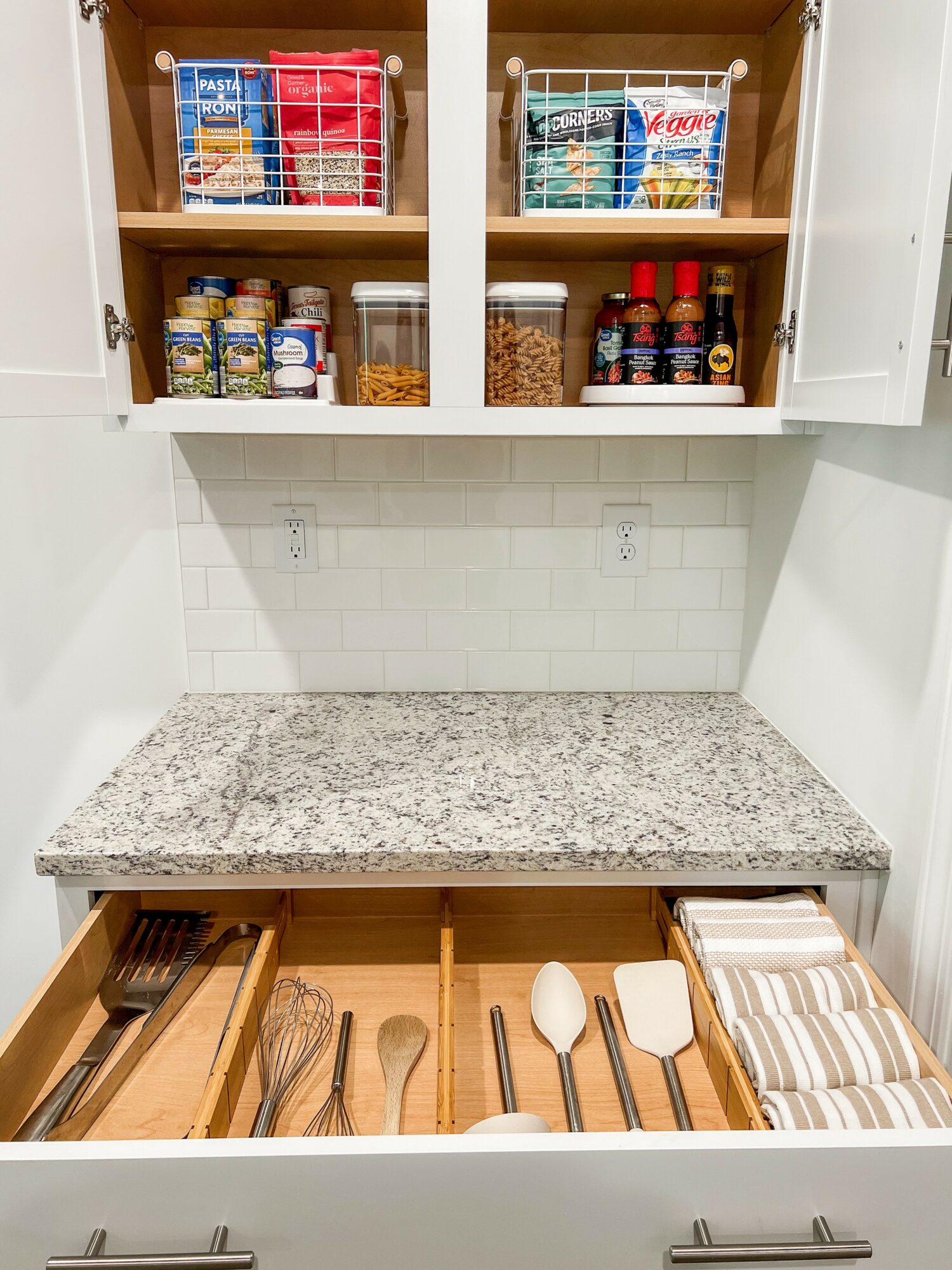 Open kitchen cabinet with pantry items on shelves and utensils in drawer, granite countertop, white tiled backsplash, and white walls.