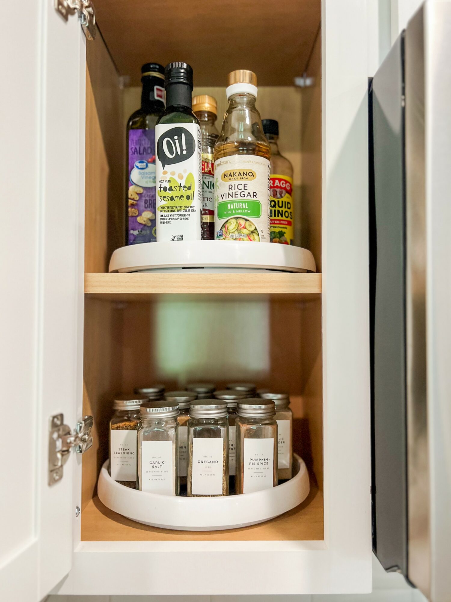 Open kitchen cabinet with bottles of oil and vinegar on two white circular trays, one on each shelf.