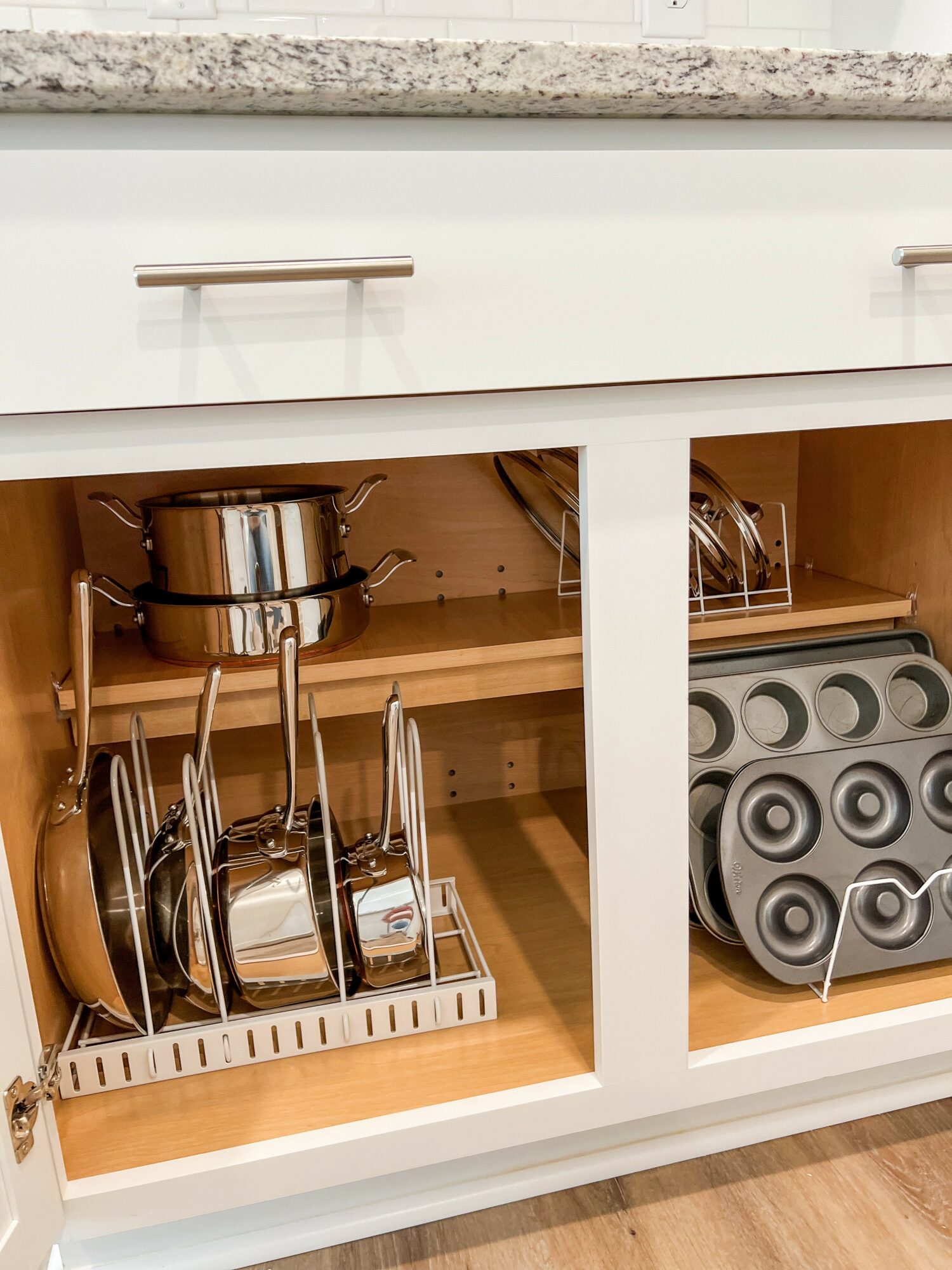 Open cabinet with pots, pans, and lids inside, organized on racks and shelves, with a granite countertop above.
