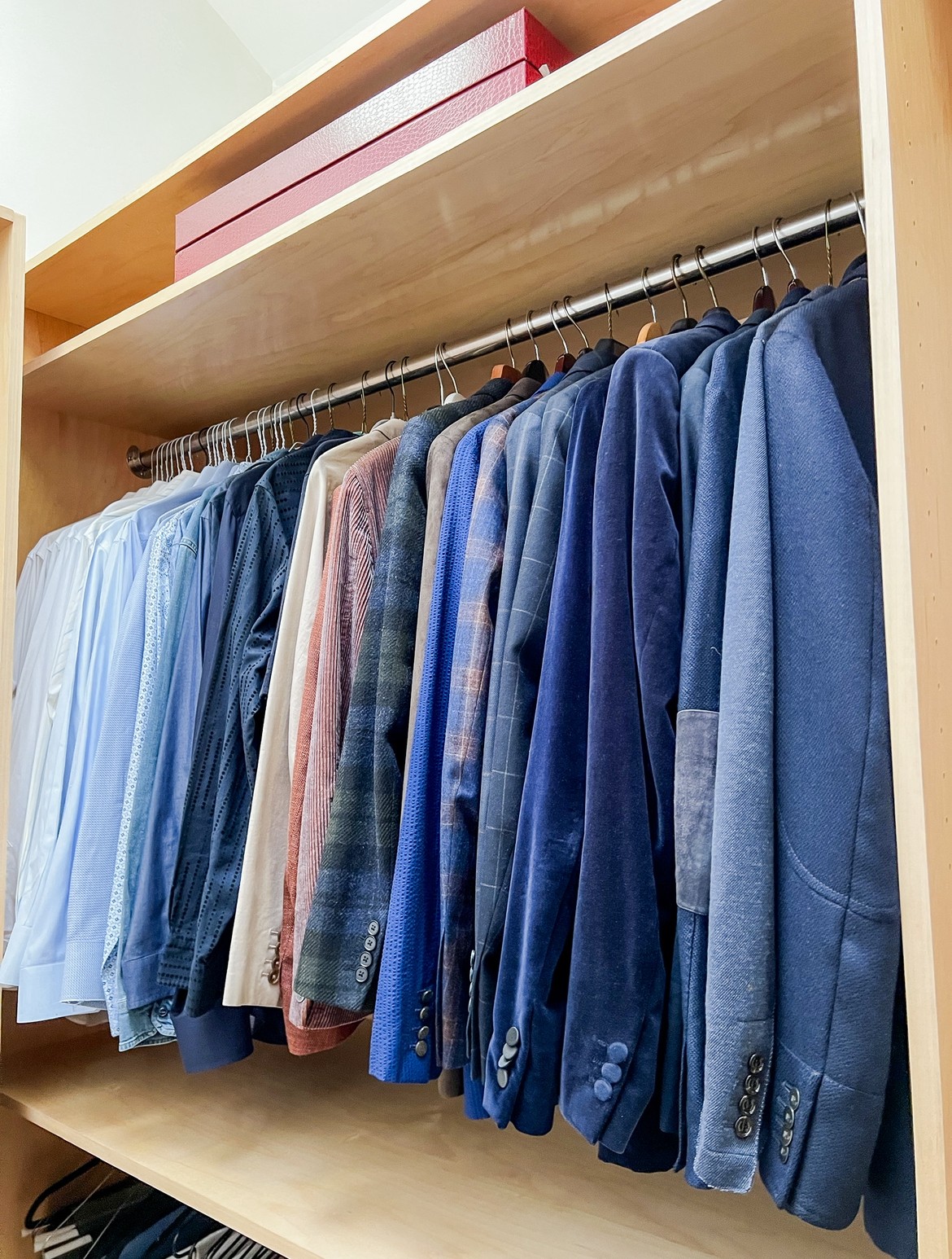 Closet with hanging shirts and jackets in various shades of blue and plaid patterns, wooden shelf above, pink box on top.